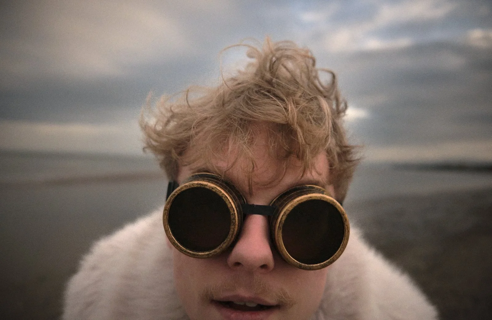 Close-up of a person with curly blond hair wearing vintage-style goggles, outdoors near water with a cloudy sky in the background.