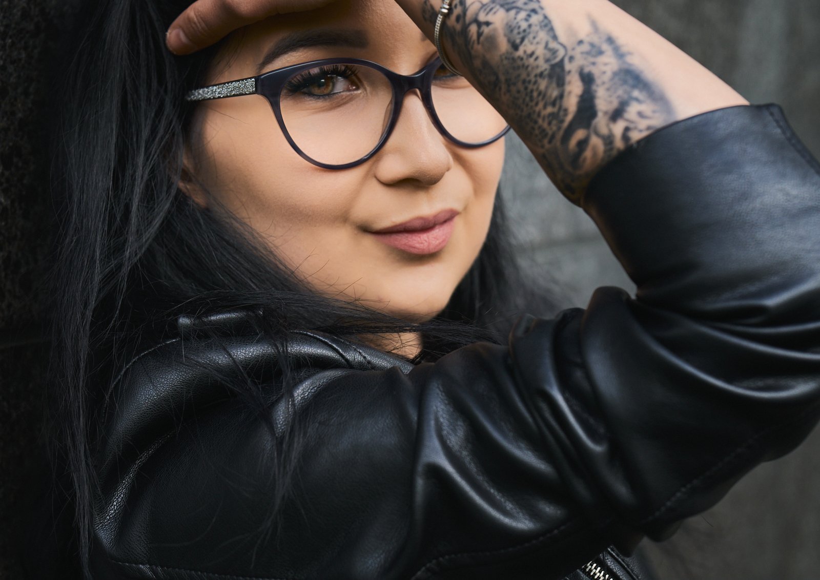 Close-up of a woman with long black hair, wearing glasses and a black leather jacket, resting her arm on her forehead and smiling gently.