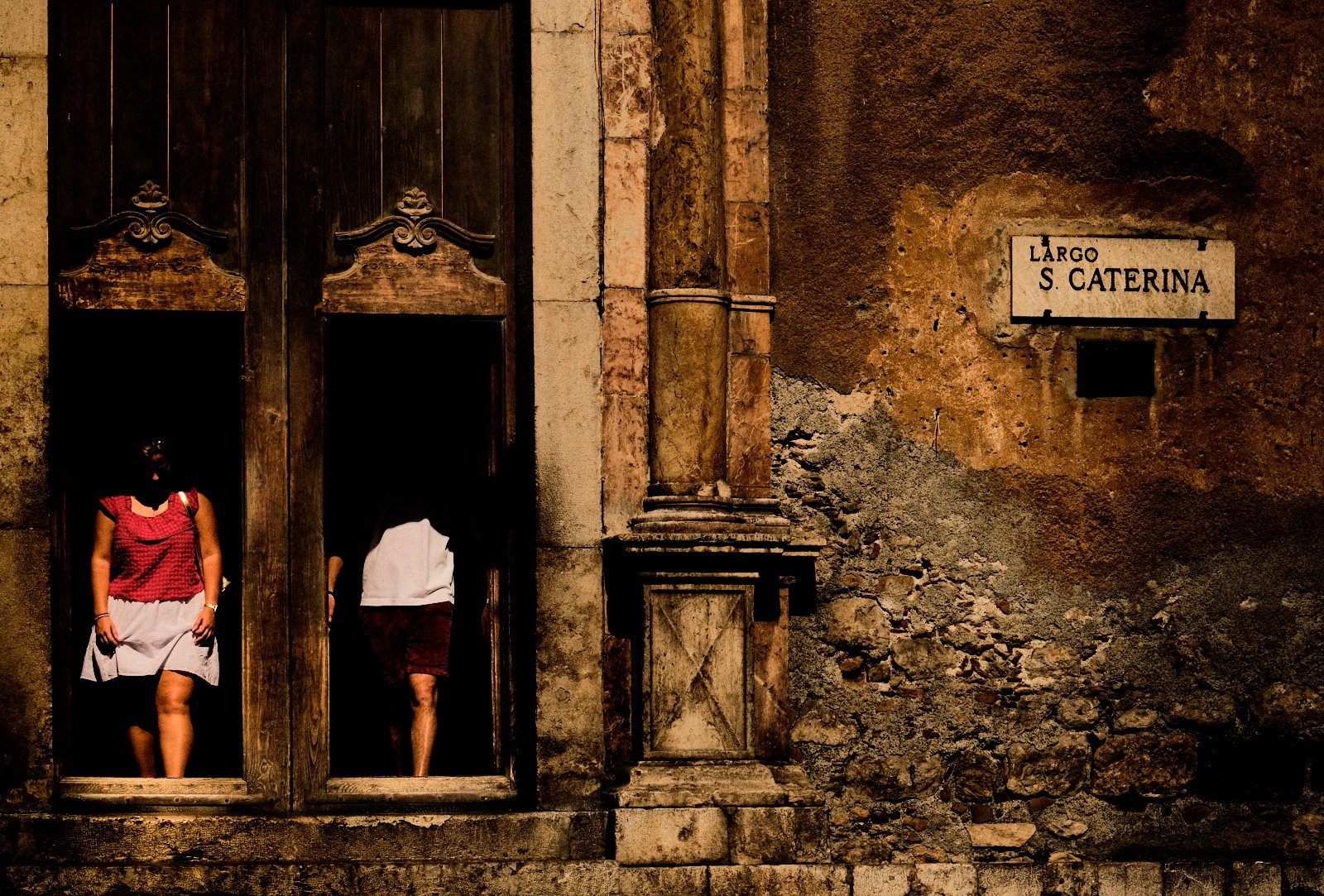 Two faceless figures in the carved wooden doorway of a church beside a weathered plaster wall, Largo Santa Caterina, Taormina — Provincia Malinconica by Tom Imio