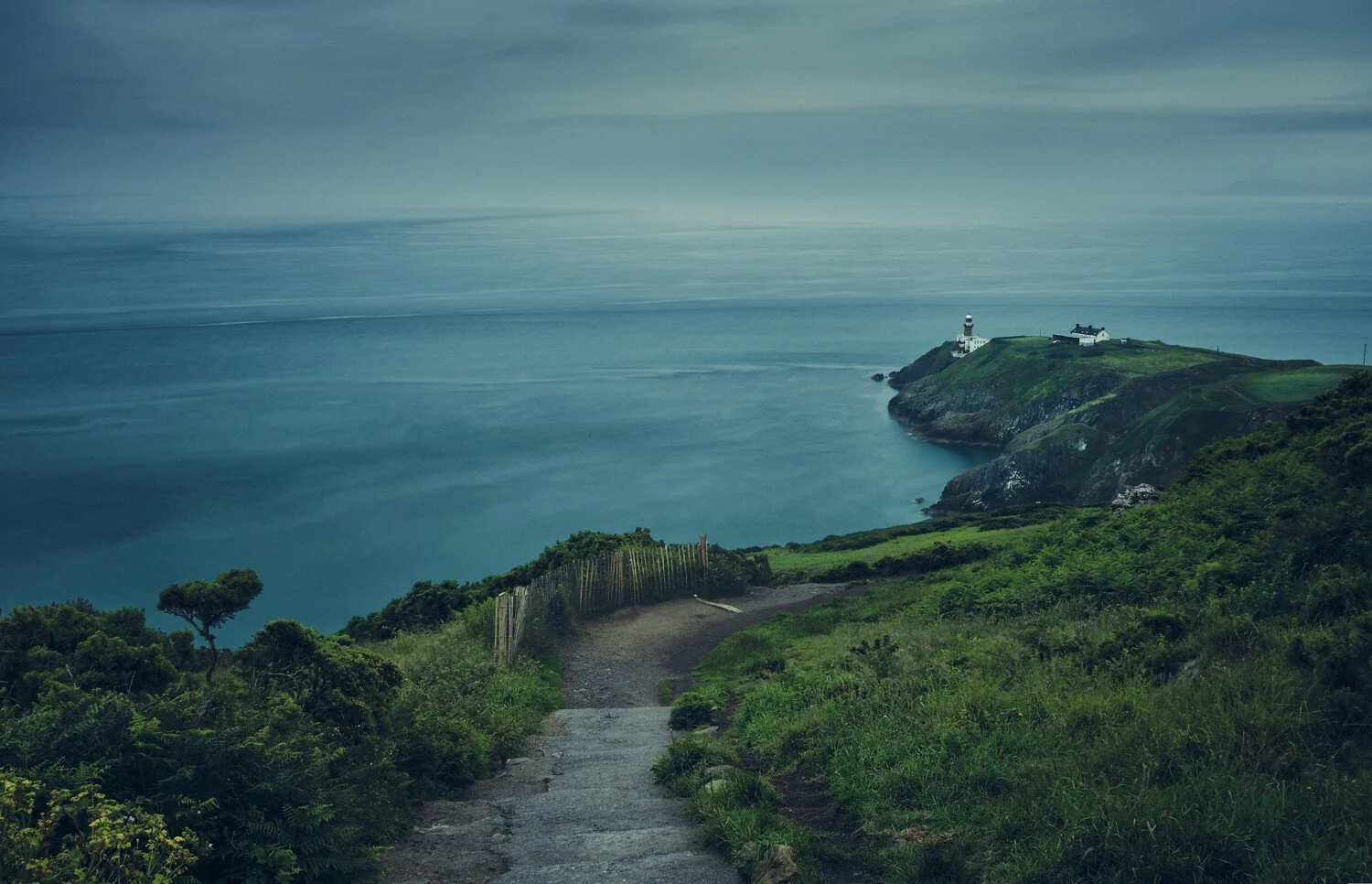 A scenic coastal landscape with a path leading to a lighthouse on a green hillside overlooking the ocean under a cloudy sky.