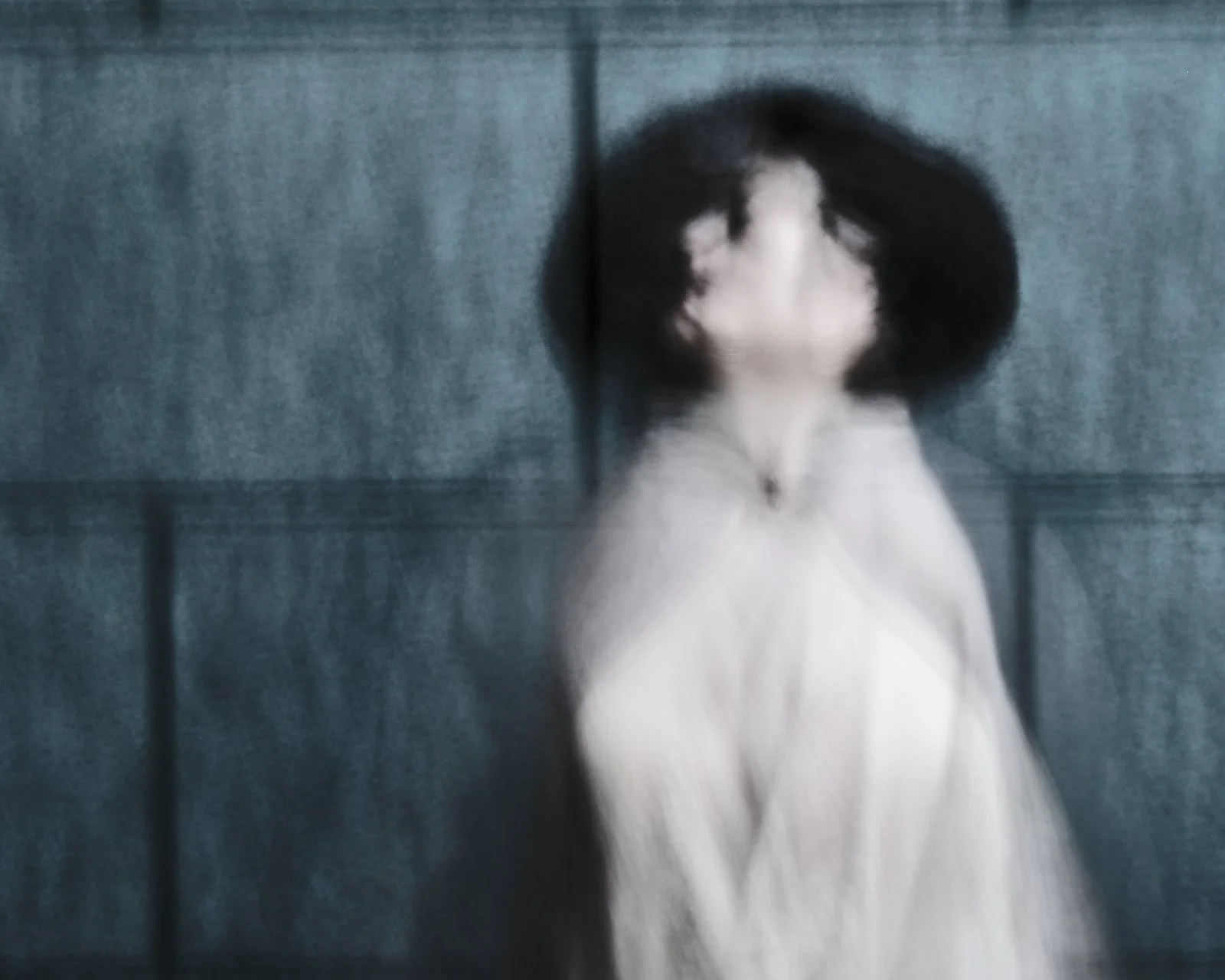 Blurred figure of a woman in a pale garment against a grey concrete wall, captured with intentional camera movement
