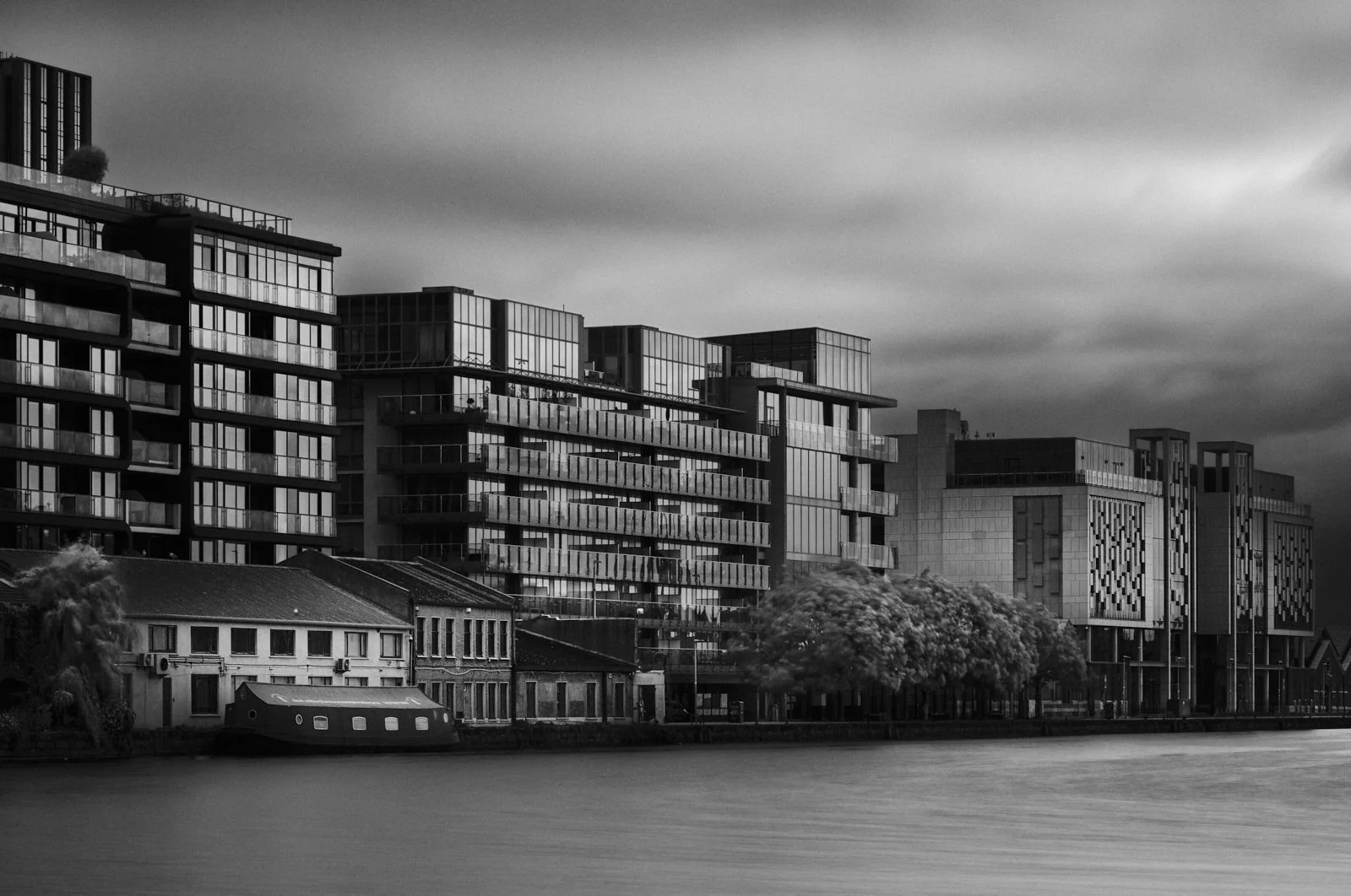 Black and white photo of modern multi-story apartment buildings with glass facades along a waterway, with an older row of houses in the foreground and cloudy sky overhead.