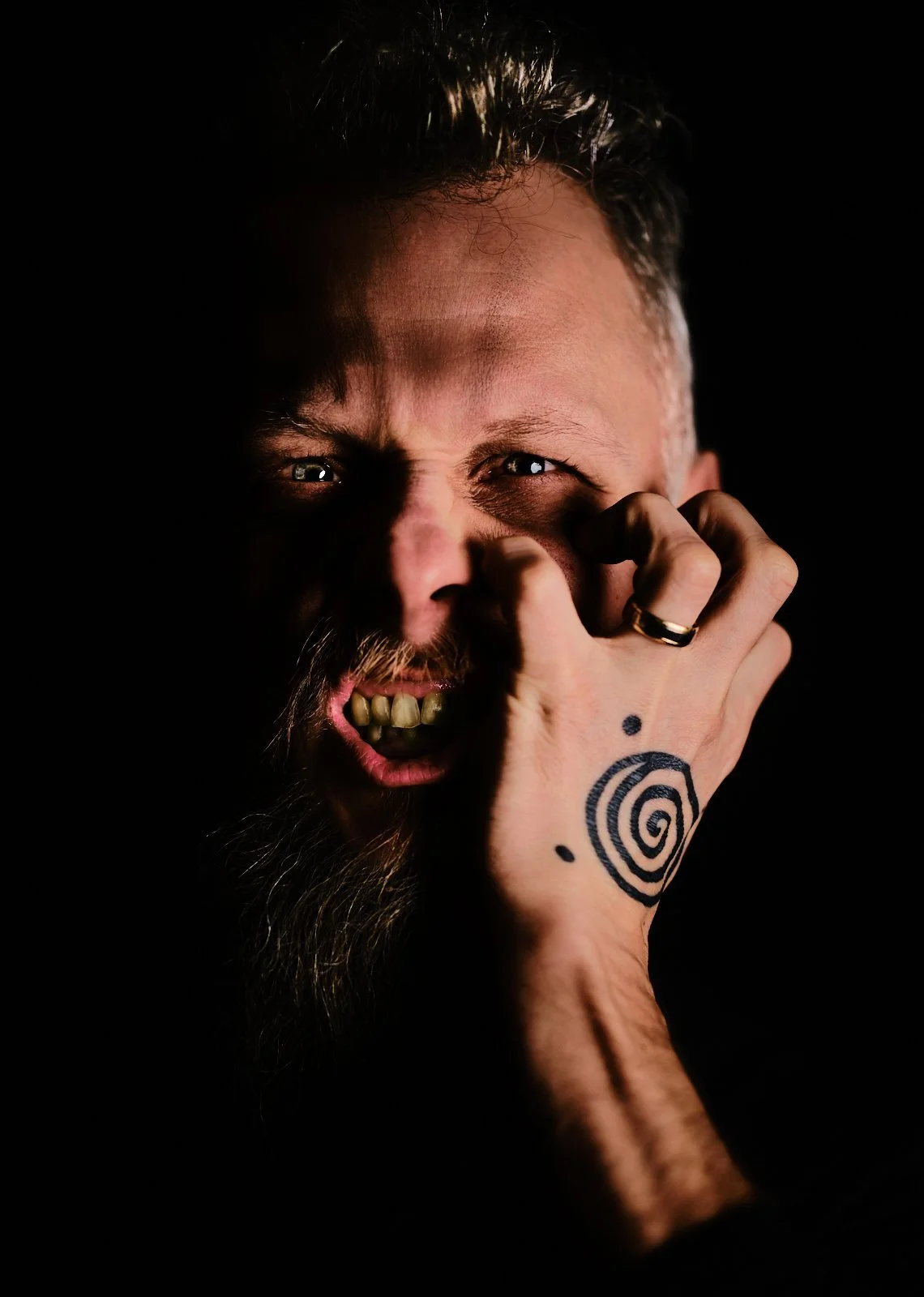 A man with a beard and tattoos on his hand appears to be in a state of anger or frustration, with his teeth bared and eyes wide open. Someone's hand with a tattoo on the thumb is gripping the man's face.