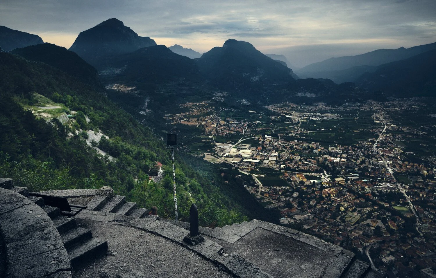 A mountain landscape view from a terrace shows a village in the valley, surrounded by steep green hills and distant mountains under cloudy skies.