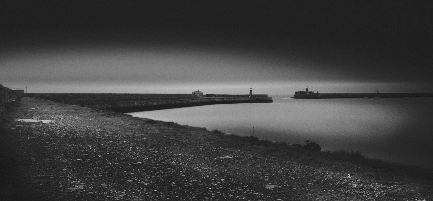 Black and white night scene of a harbor with a curved breakwater, lighthouse, and calm water.