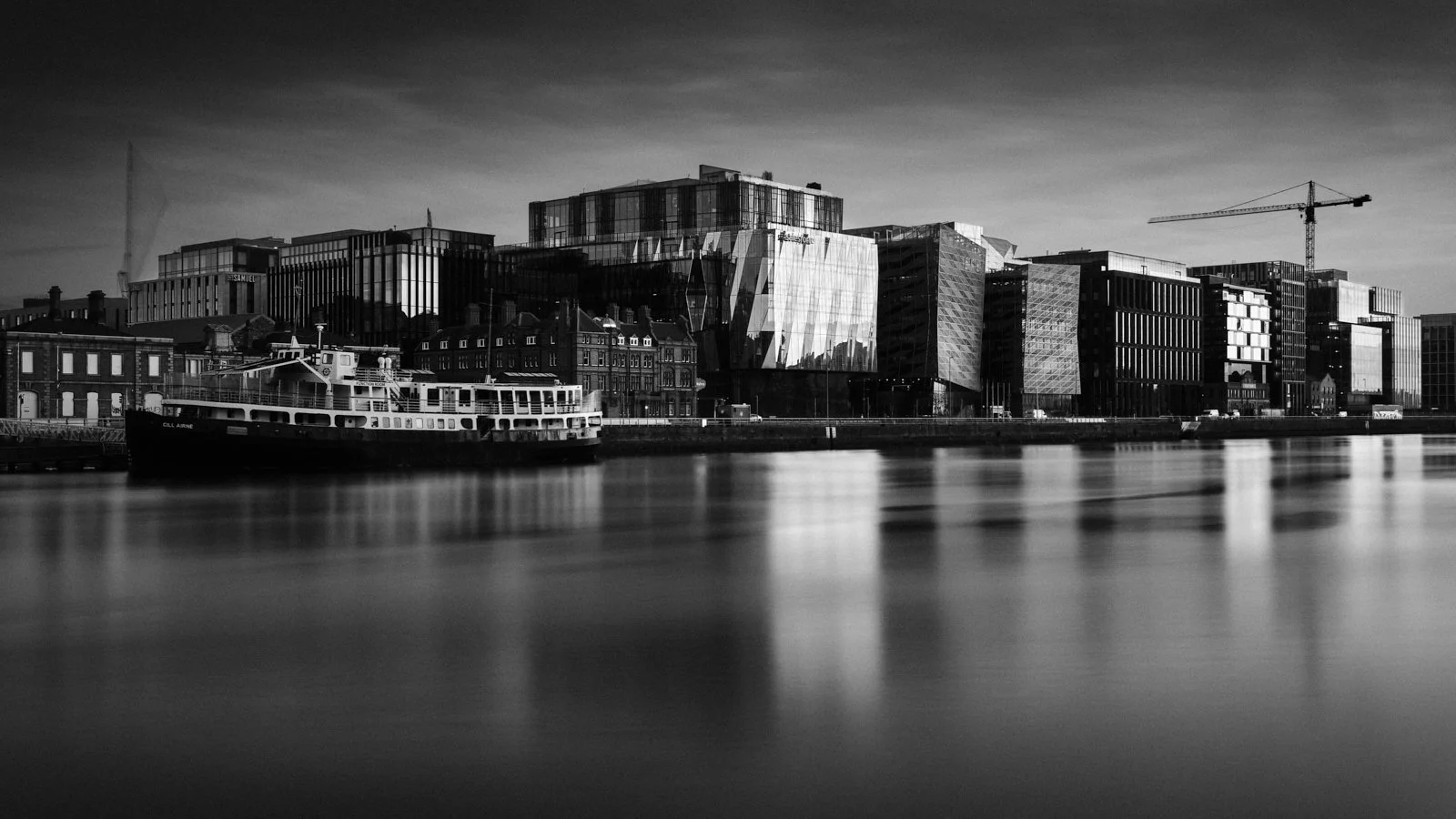 Black and white photo of modern city buildings along a waterway with a boat docked at the waterfront.
