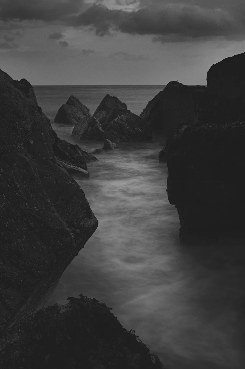 Black and white photograph of a rocky seascape with large dark rocks in the foreground and a cloudy sky overhead.