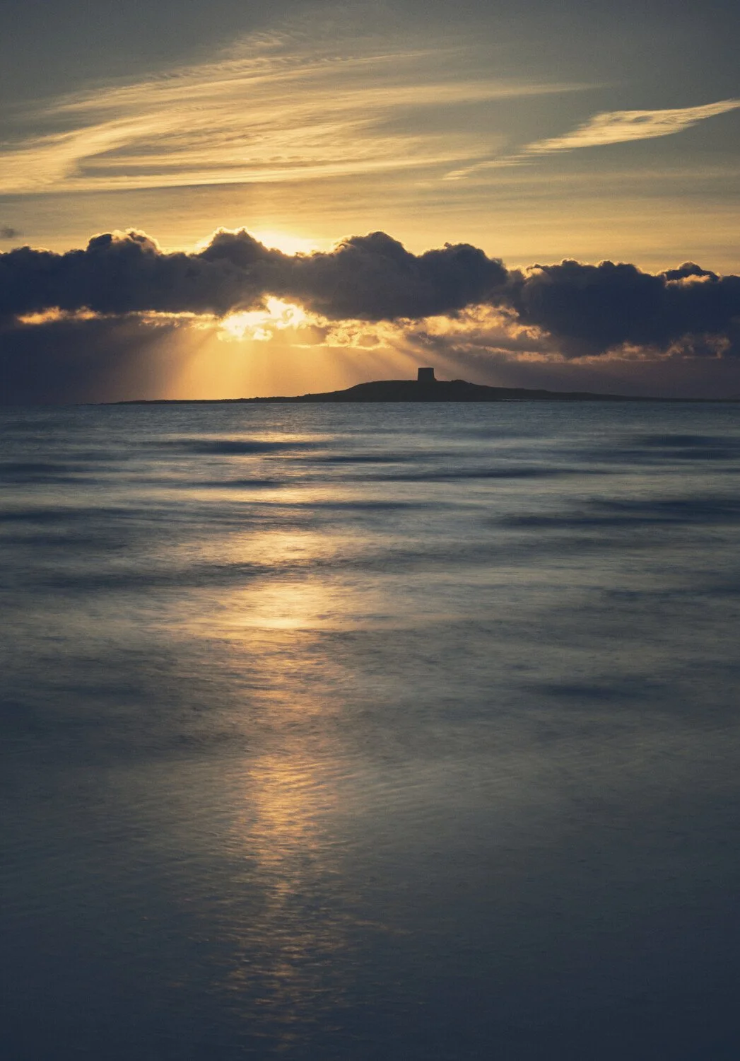 Sunset over water with clouds and a distant island with a small tower or building.
