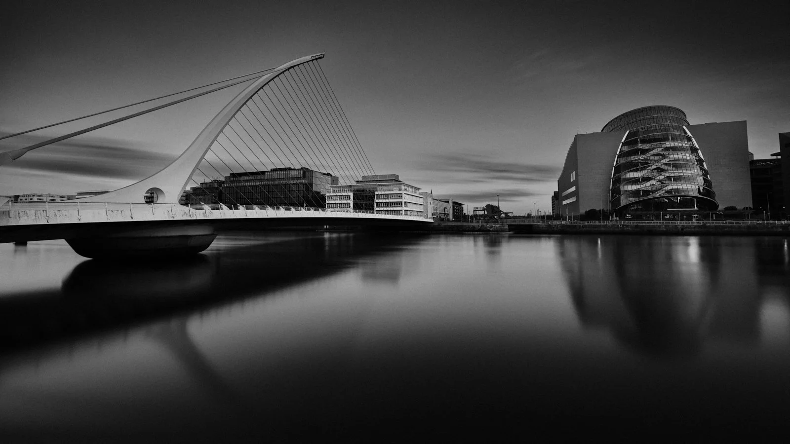 Black and white photo of the Dublin docklands including the Samuel Beckett Bridge with its distinctive harp shape, and the cylindrical shaped modern building at the Convention Centre Dublin, reflected on calm water.