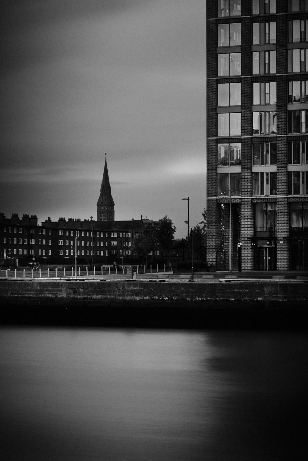 A black and white cityscape featuring a modern apartment building on the right and a church steeple in the background, with a water body in the foreground.