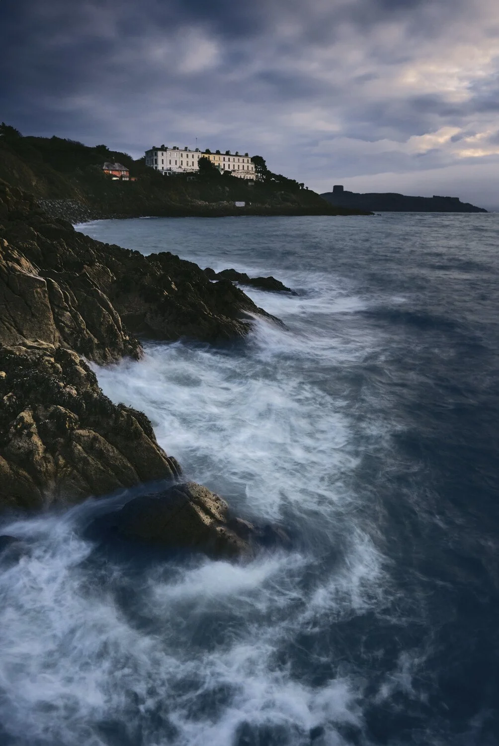 Coastal scene with rocky shoreline and waves crashing, in front of a large white building on a hill, under a cloudy sky.