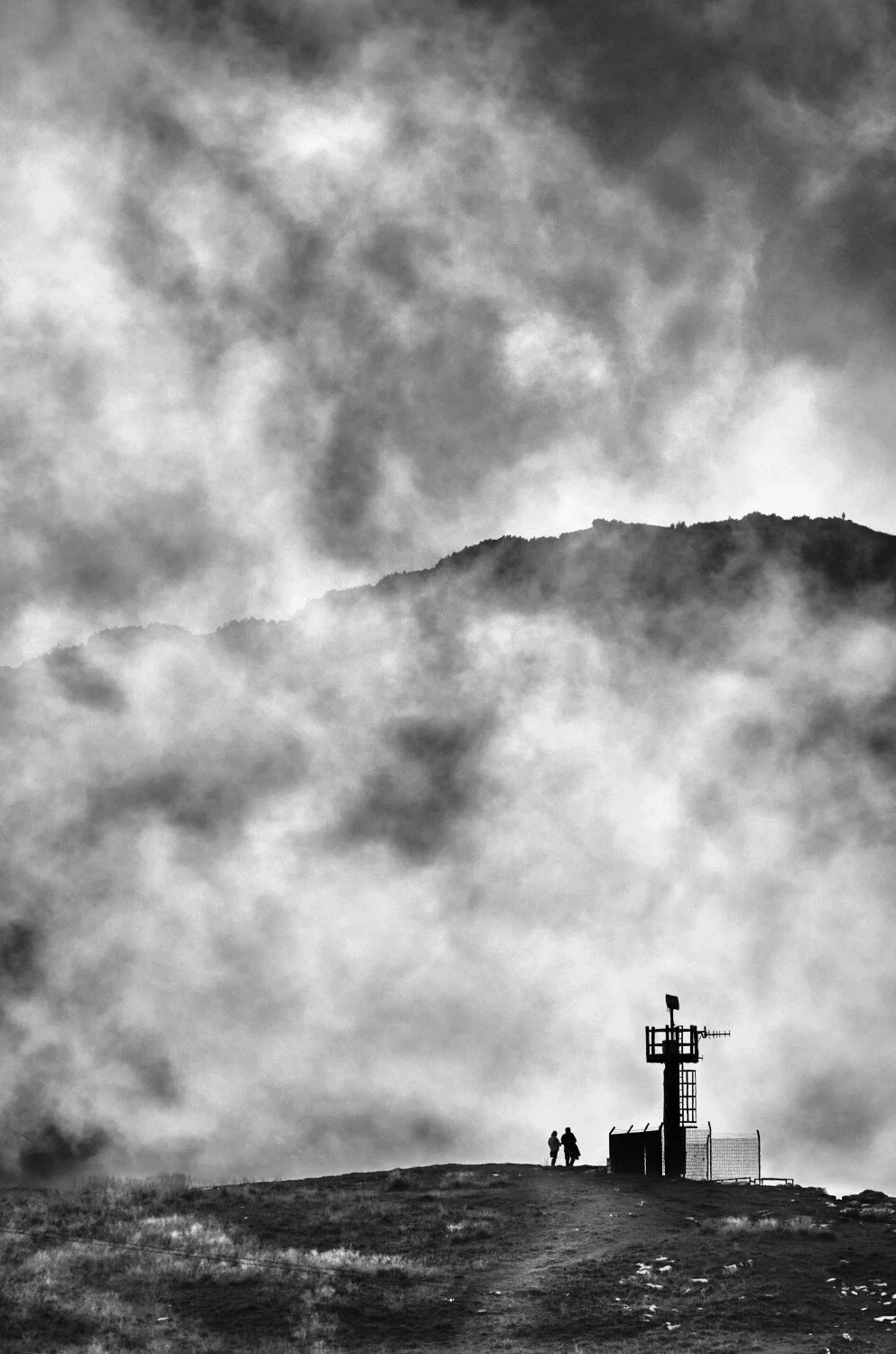 Two people standing on a hilltop near a small communications tower, with mountain and cloudy sky in the background.