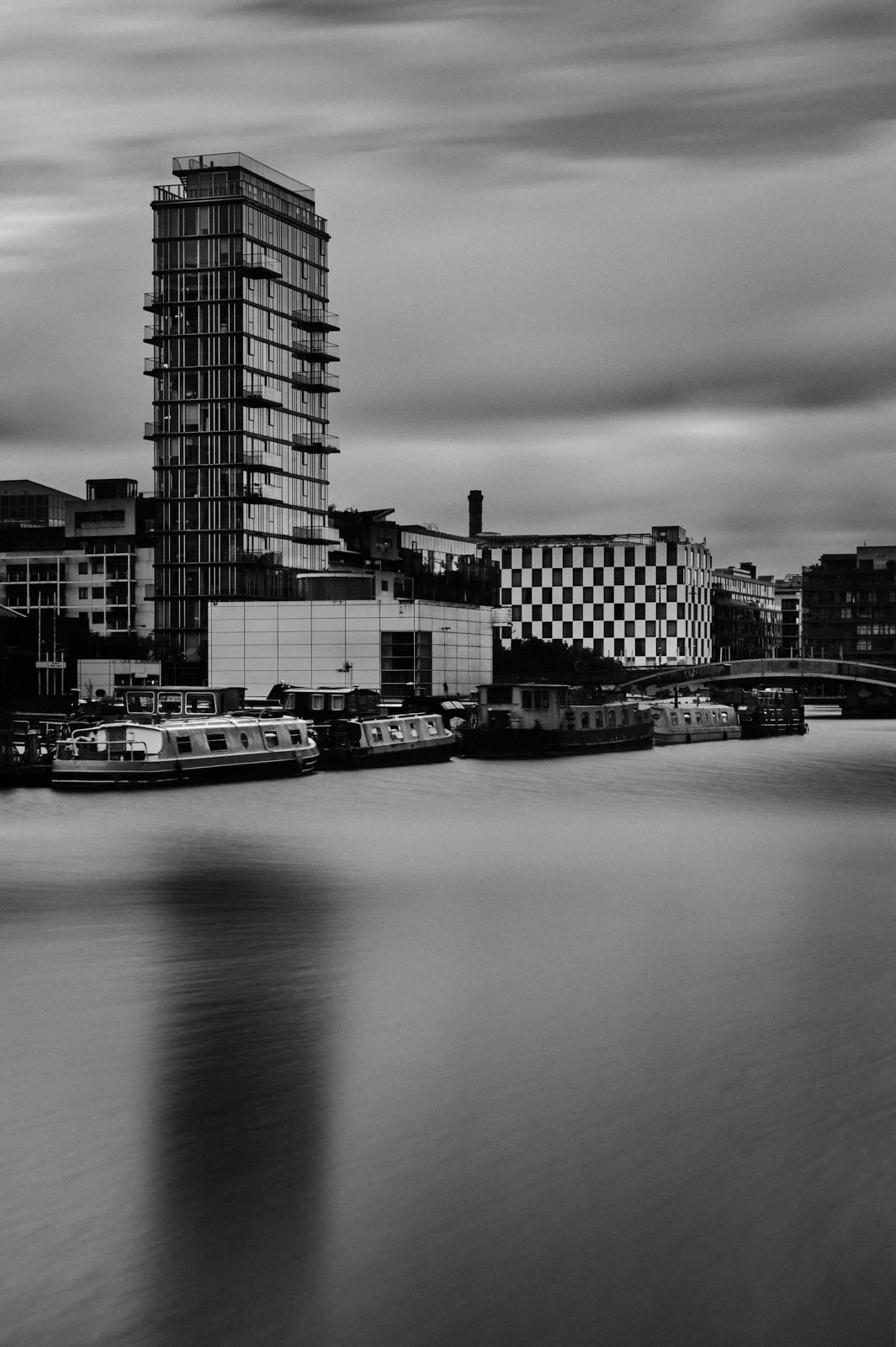 Black and white photo of modern cityscape with tall building and boats on water.