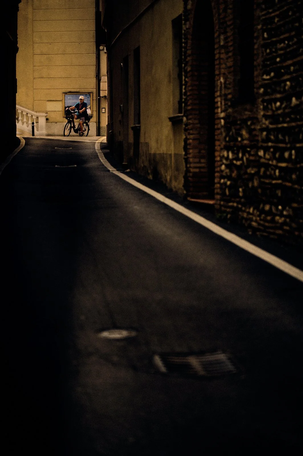 Cyclist framed in a pocket of warm light at the end of a dark, curving alley in Desenzano del Garda, Italy — Provincia Malinconica, fine art street photograph by Tom Imio