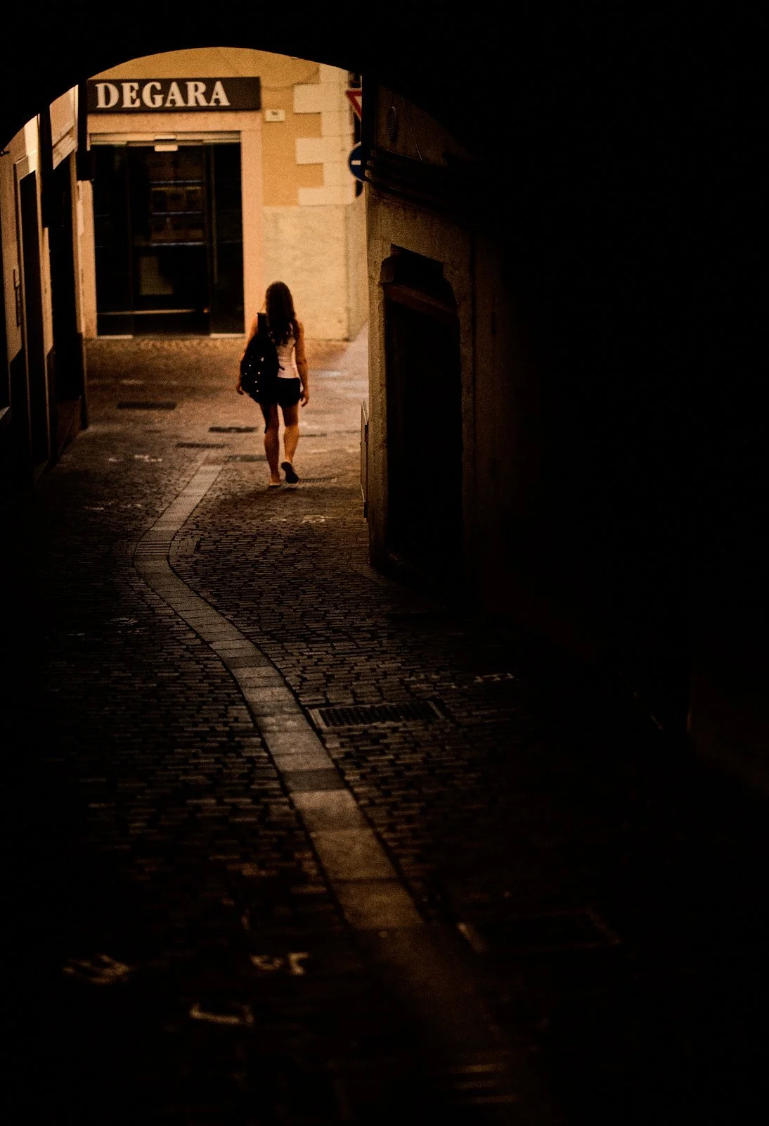 Woman with a backpack walking away through a dark stone archway toward a sunlit alley in Riva del Garda, Italy — Provincia Malinconica by Tom Imio