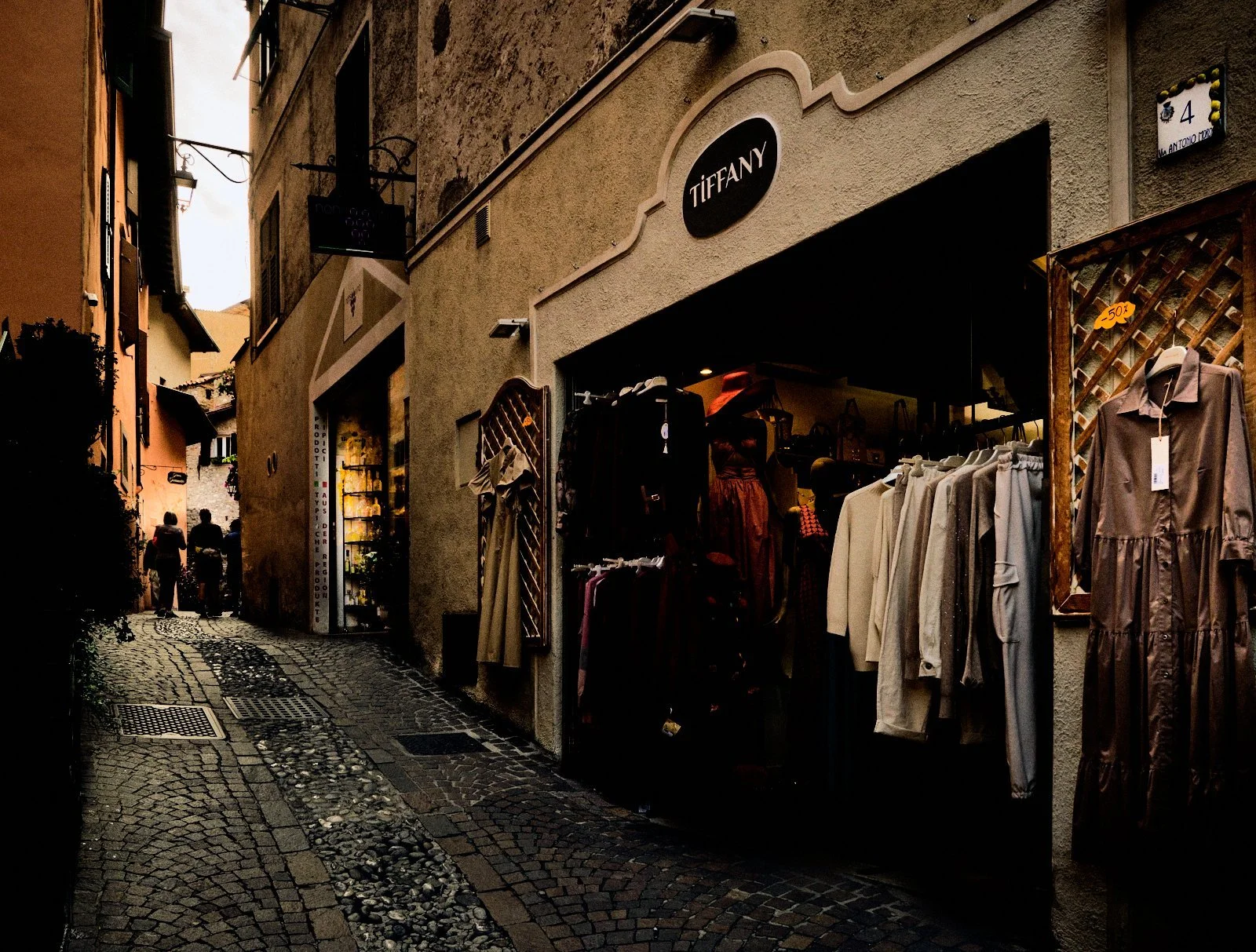 Fashion boutique spilling clothes racks into a historic cobbled alley in Limone del Garda, figures dissolving into the light beyond — Provincia Malinconica by Tom Imio