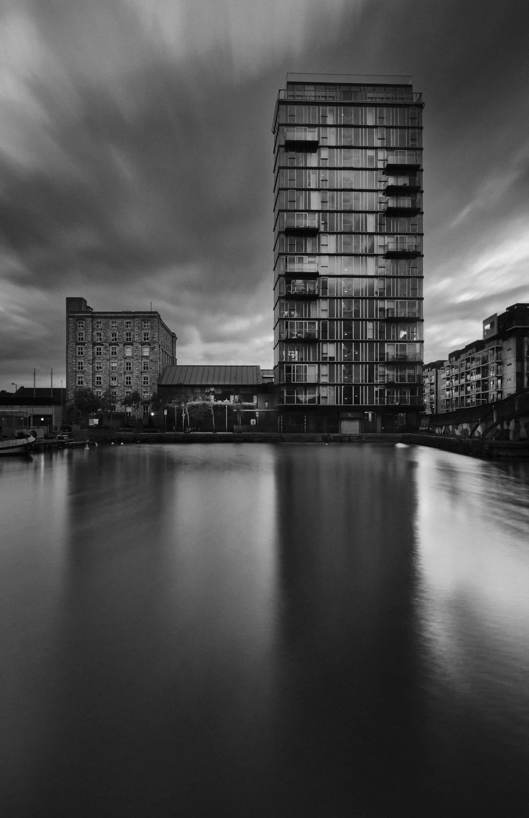Black and white photograph of a tall modern apartment building with glass balconies, reflected in a calm body of water, with a cloudy sky above.