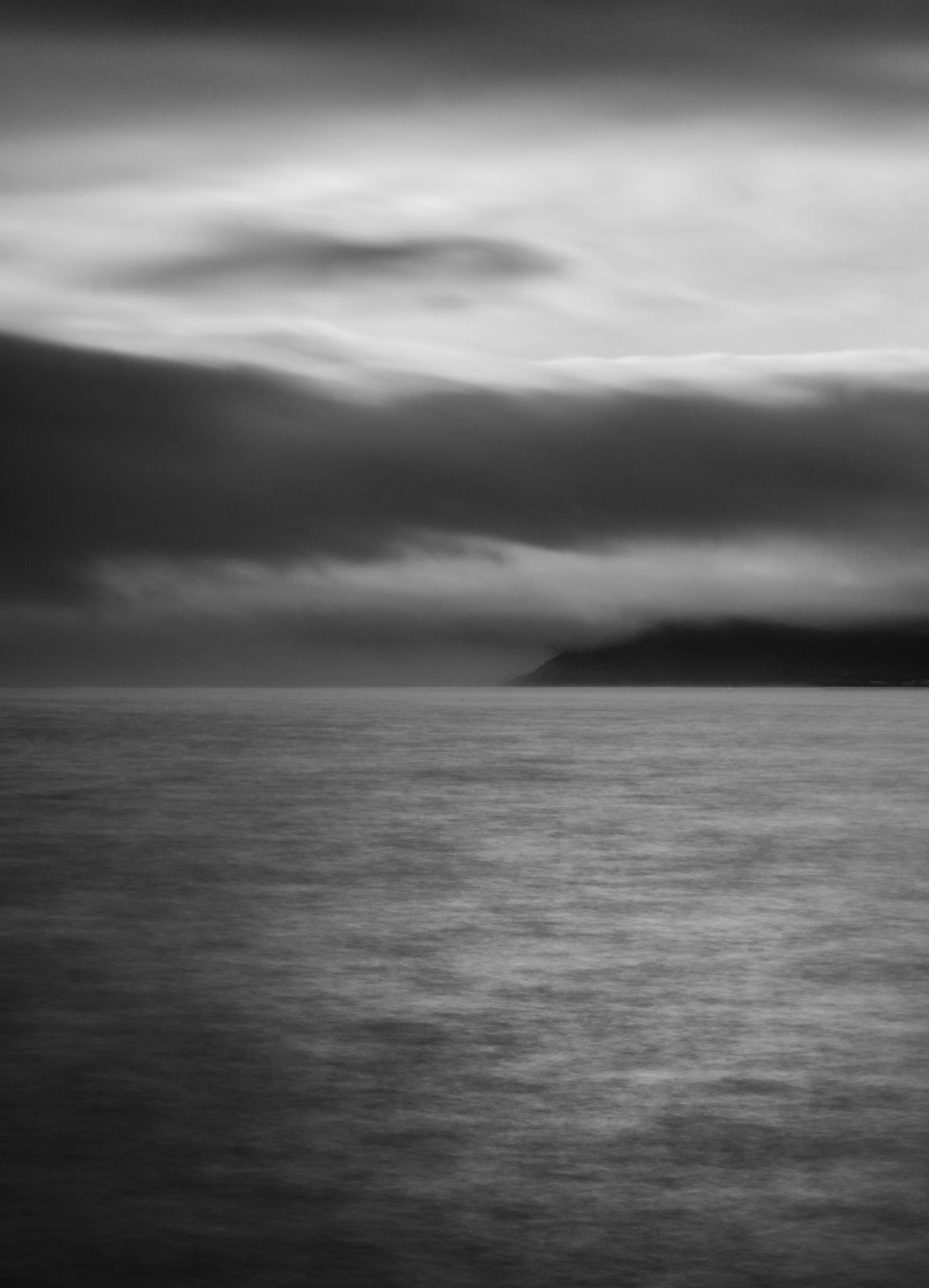 Black and white photo of a calm ocean with a cloudy sky, distant landmass on the horizon.