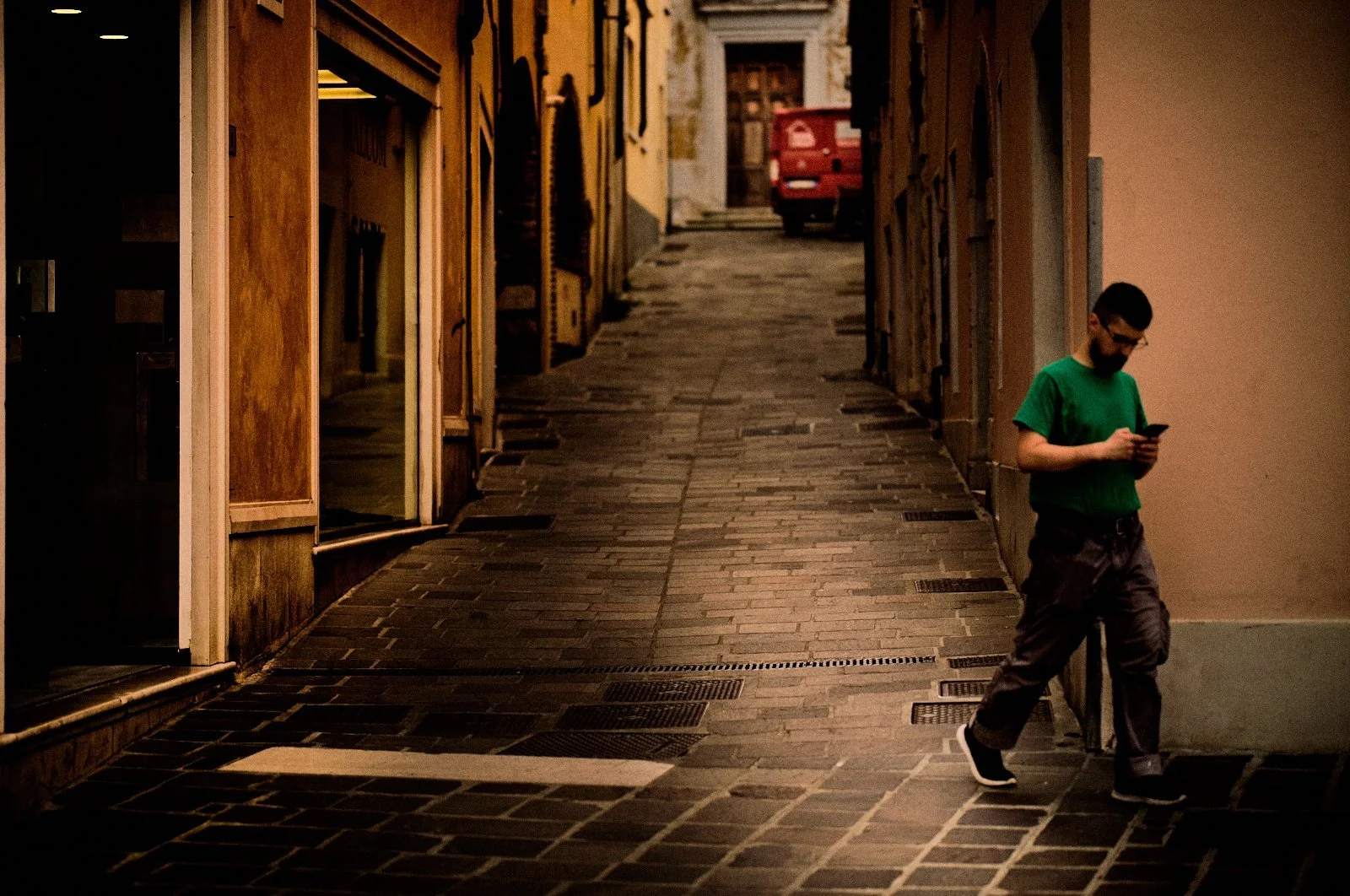 Man in a green t-shirt absorbed in his phone on an empty cobbled street at dusk in Desenzano del Garda, Italy — Provincia Malinconica by Tom Imio