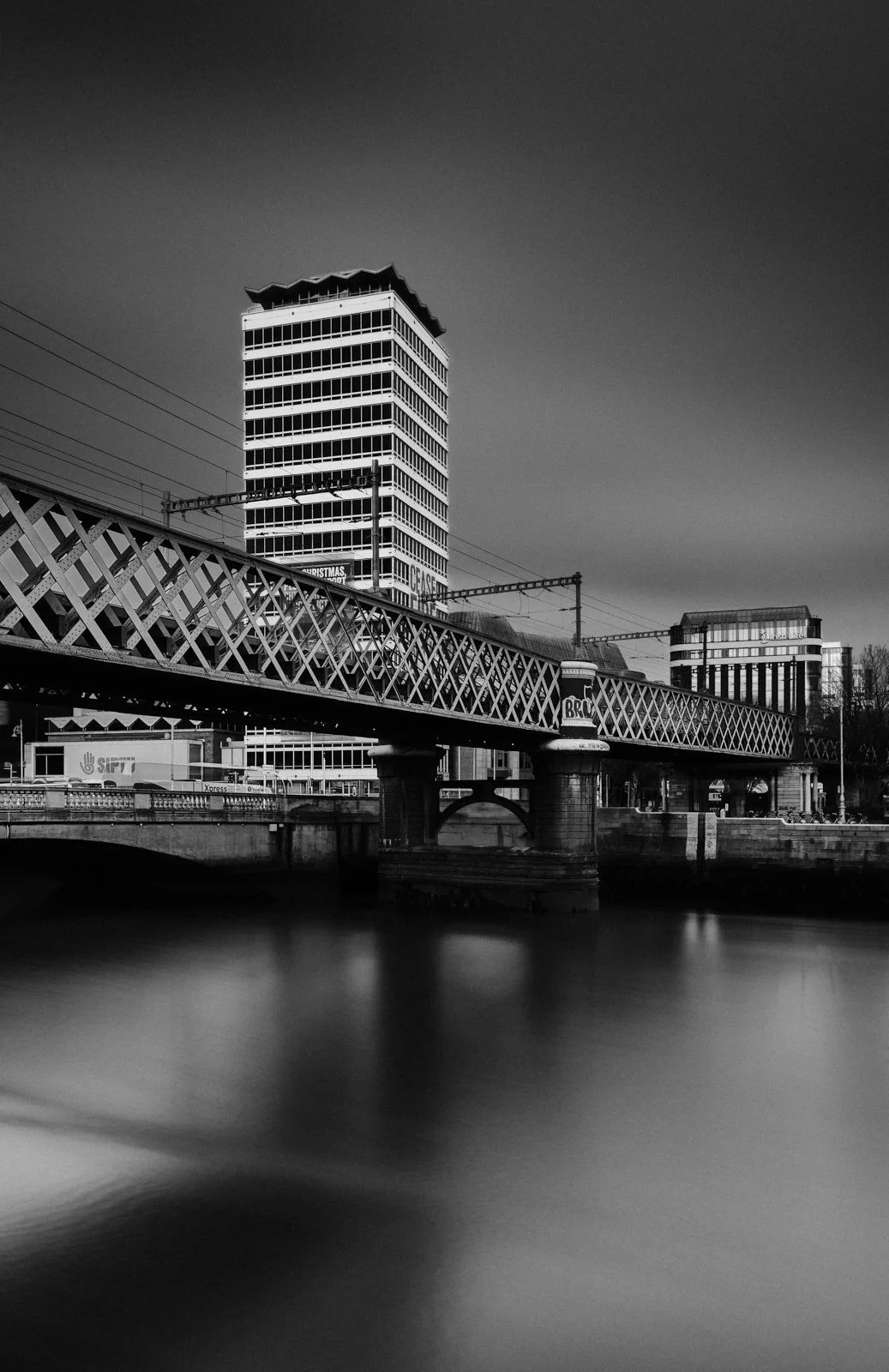 Black and white photo of urban buildings and a bridge over water, with a tall modern skyscraper in the background.
