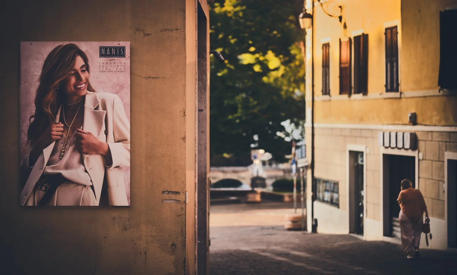 Luxury fashion poster on a weathered wall, while an elderly woman walks away down a golden-lit Italian street