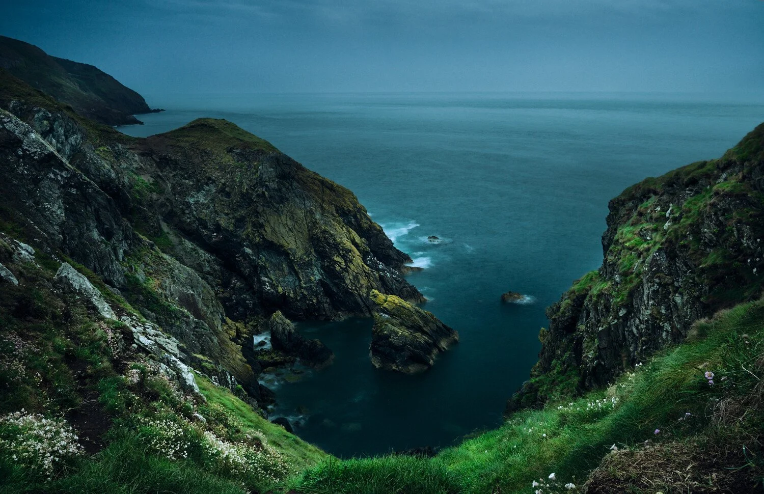 Coastal landscape with steep green and rocky cliffs overlooking a calm blue ocean, under a cloudy sky.