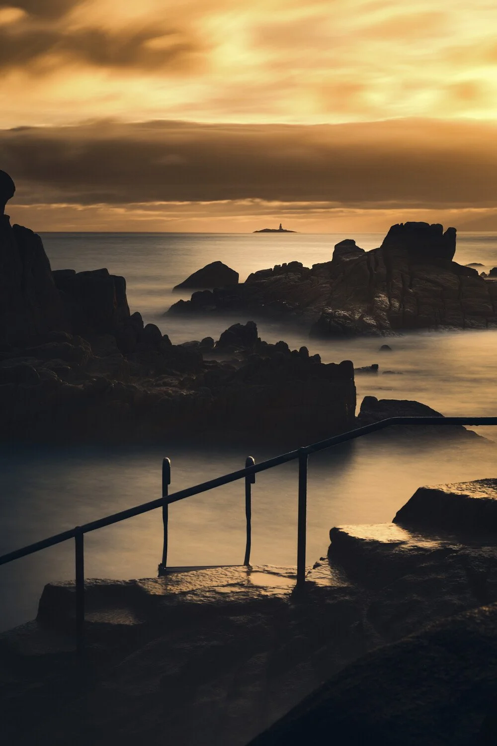 A rocky shoreline at sunset with a railing in the foreground, calm ocean waves, clouds, and a small island with a lighthouse in the distance under a dramatic orange sky.