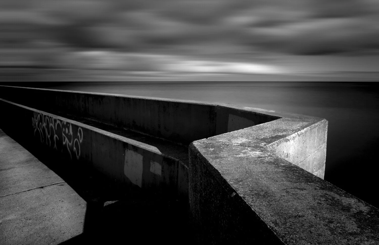 Black and white photo of a concrete seawall or barrier along the shoreline with graffiti on one side, under a cloudy sky.