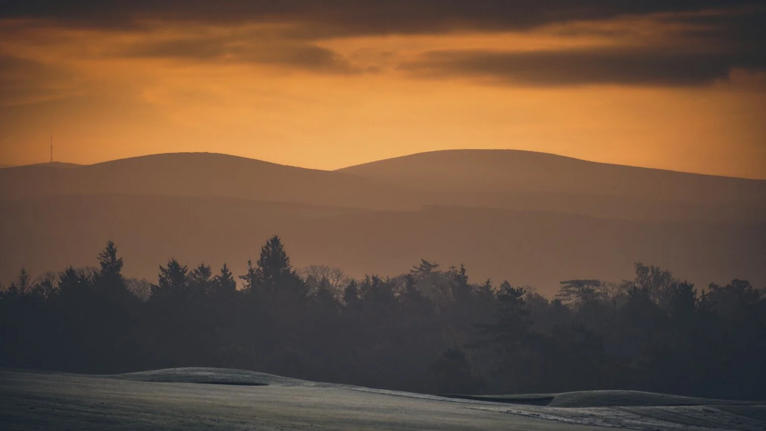 Landscape of rolling hills and trees at sunset with a partly cloudy sky.