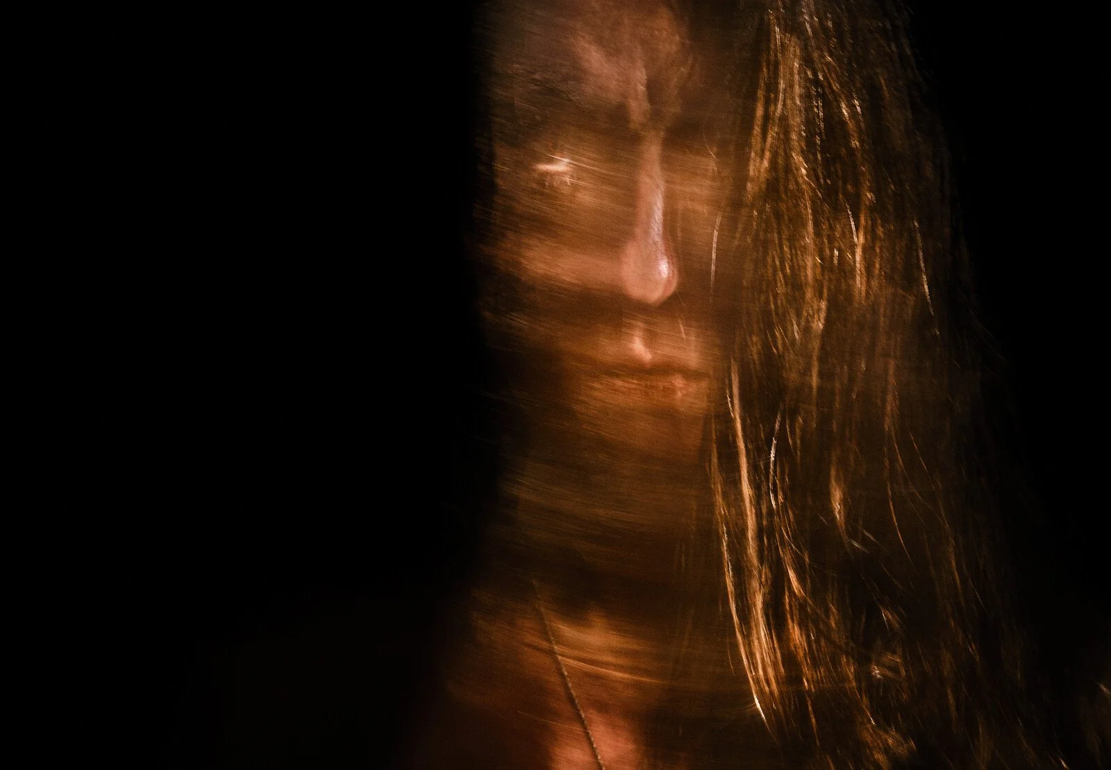 A person's face is partially illuminated with warm light, creating a moody, artistic effect with motion blur, long hair, and closed eyes.