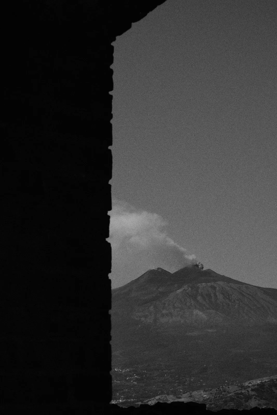 Mount Etna with a rising ash plume seen past the dark edge of a stone wall, Taormina — Whispers of Form by Tom Imio