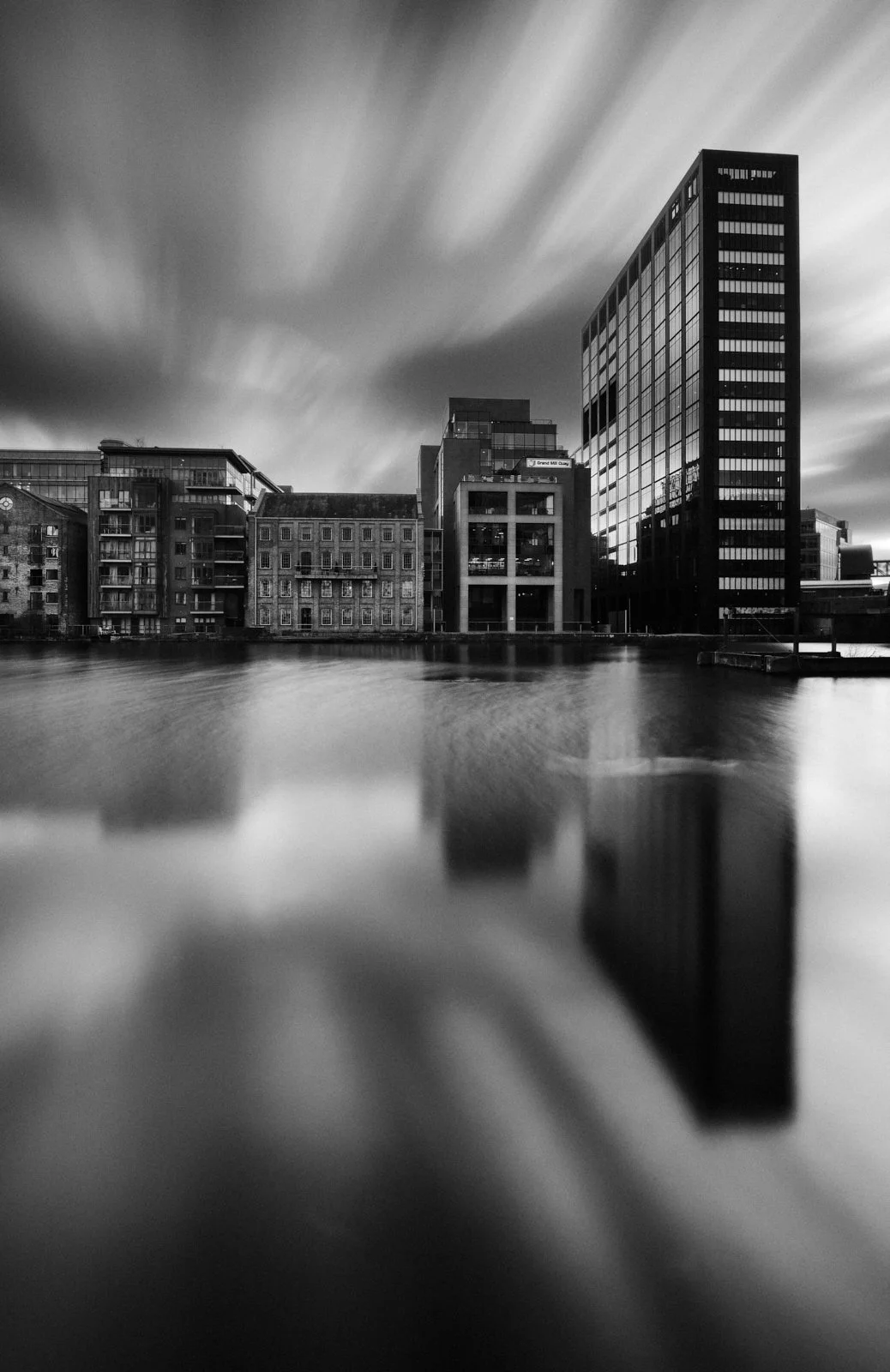Black and white photo of modern buildings along a river with reflections, under a cloudy sky with streaked clouds.