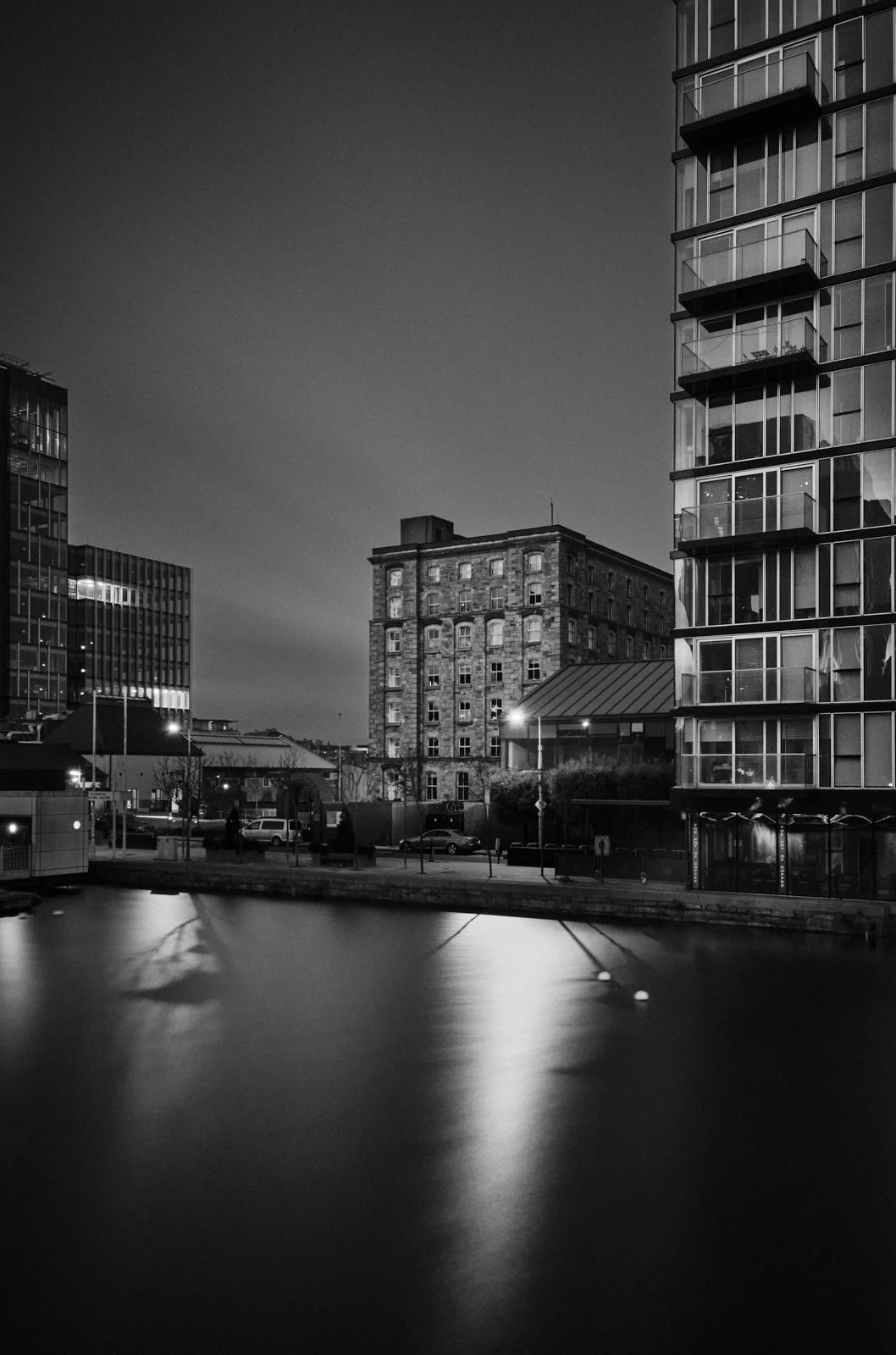 Black and white photo of city buildings at night, with water in the foreground reflecting the lights from the buildings.