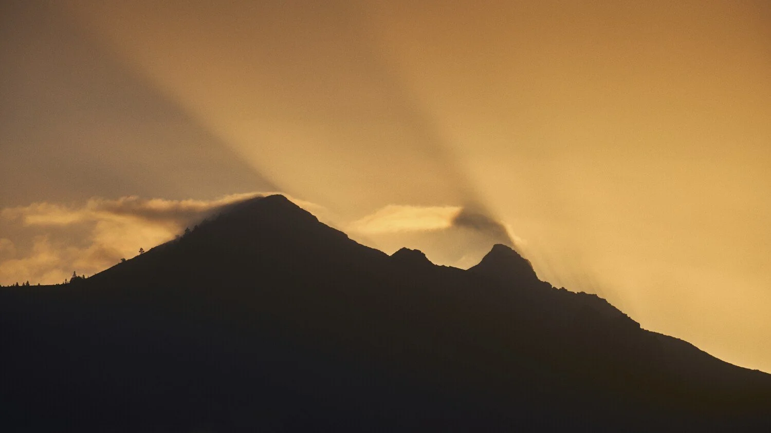 Silhouetted mountain at sunset with rays of light shining through clouds.