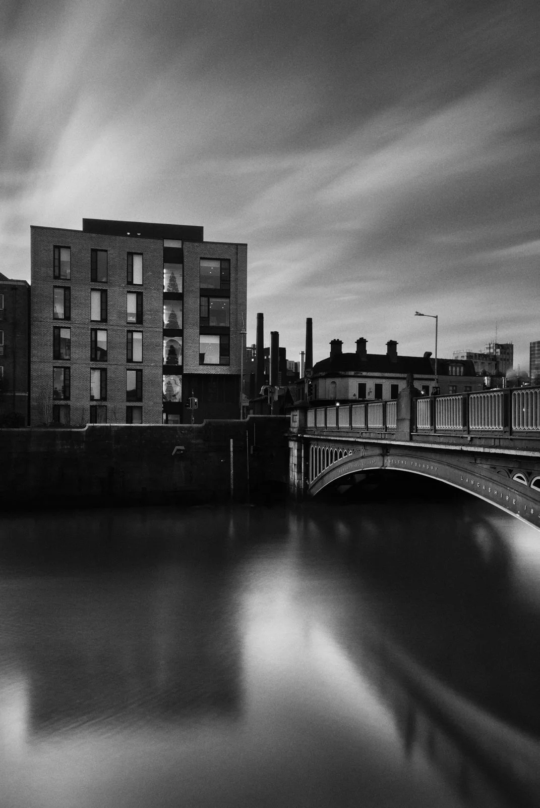 Black and white photograph of modern building and bridge over water, with a cloudy sky overhead.