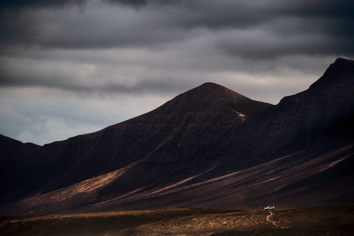 A dark, mountainous landscape with looming clouds and a solitary white vehicle on a dirt road.
