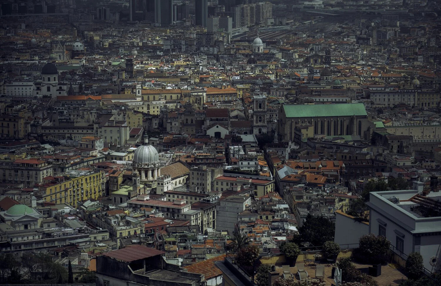 Aerial view of a densely packed cityscape with historic buildings, churches, and towers, including a prominent gray-domed church and a large cathedral with a green roof.