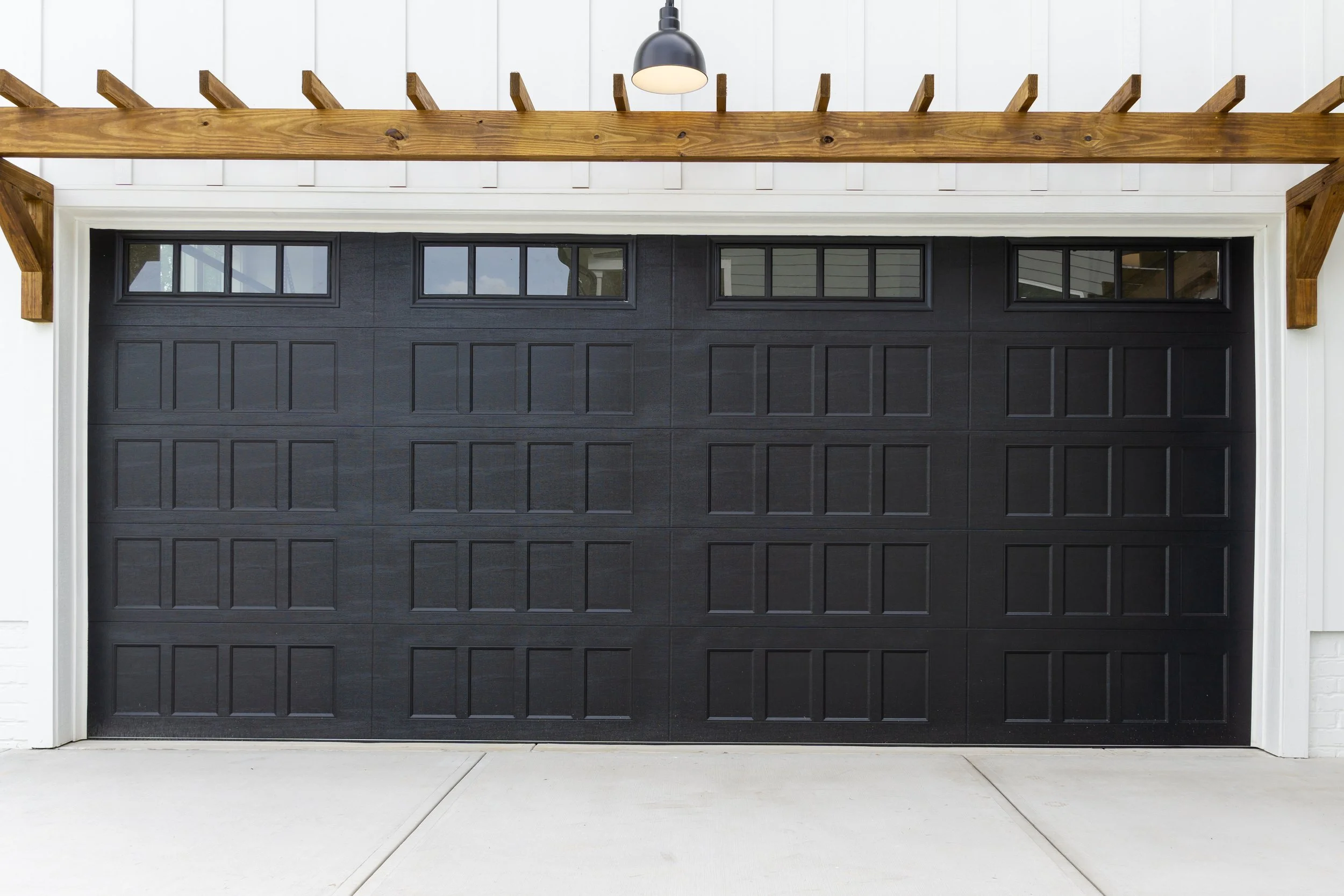 Black raised panel garage door with upper windows beneath a wooden pergola