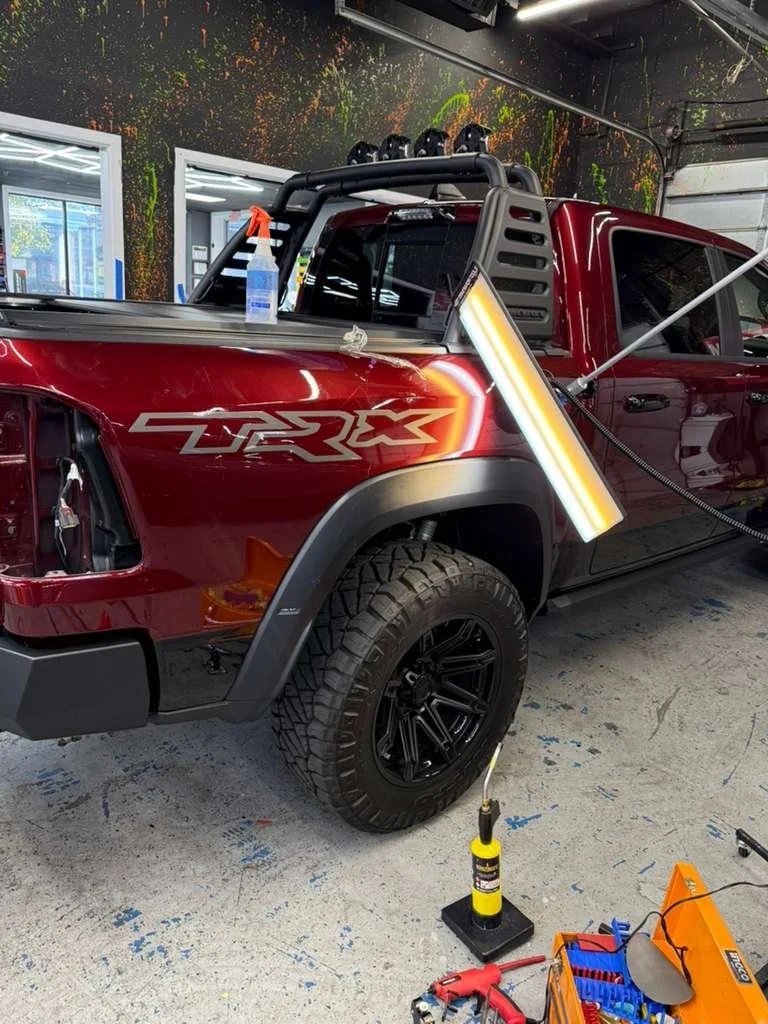 Red pickup truck with TRX and Ram logo graphics, equipped with off-road accessories like lights, a roll cage, and a front windshield protector, inside an auto workshop with tools and spray bottles nearby.