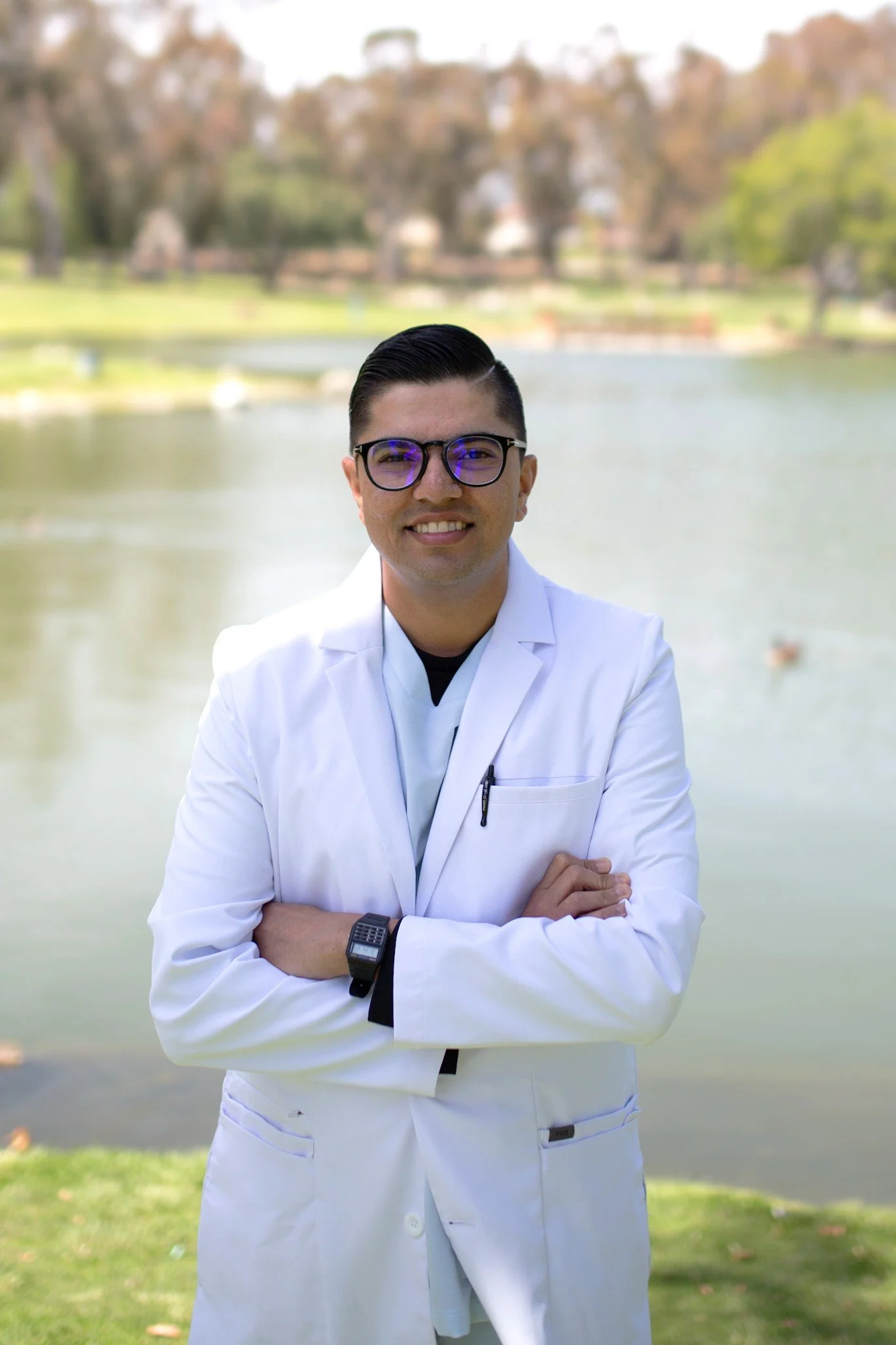 A man wearing glasses and a white lab coat standing outdoors near a lake, smiling with arms crossed.