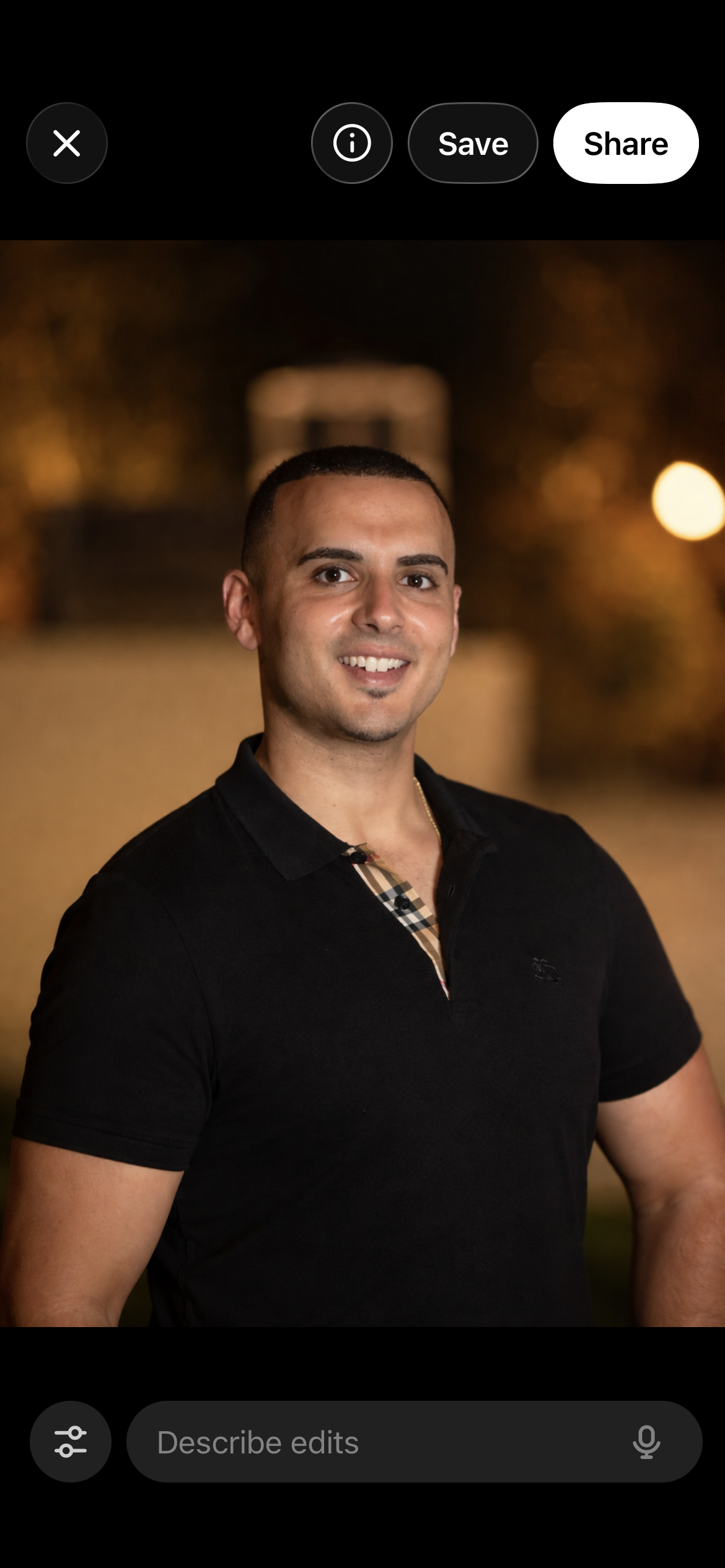A smiling man with short dark hair wearing a black polo shirt with a Burberry pattern collar, standing outdoors at night with blurred warm lights in the background.