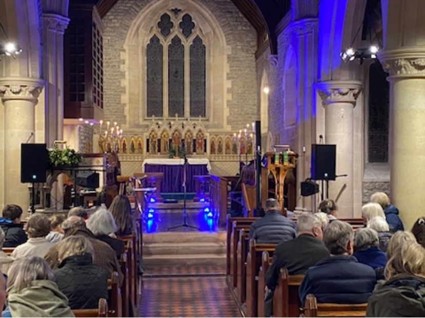 Inside of a church with people seated in pews facing an altar, with lit candles and stained glass windows in the background.