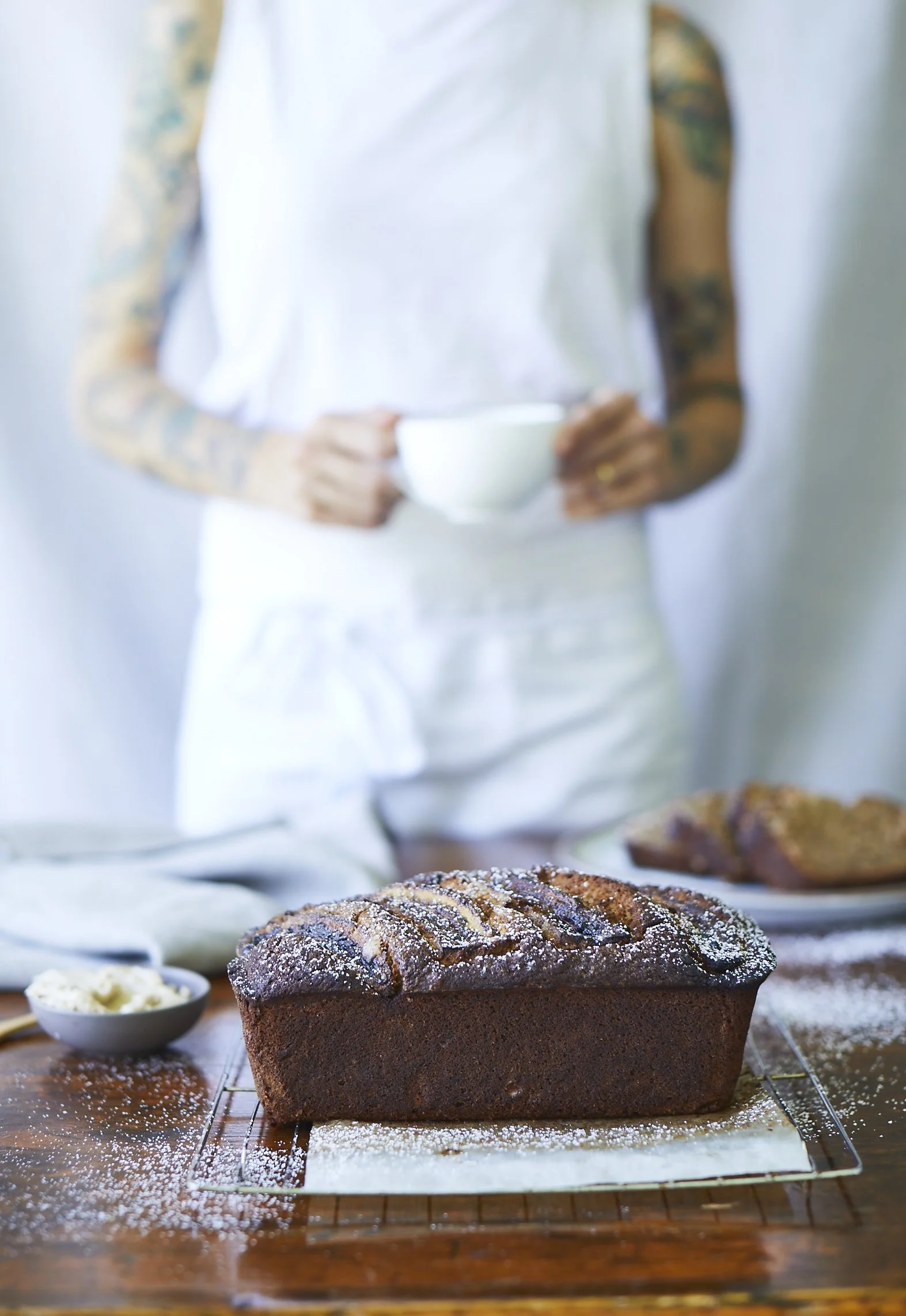 A freshly baked loaf of chocolate bread dusted with powdered sugar on a cooling rack, with a person holding a cup in the background, and various baked goods on a wooden table.