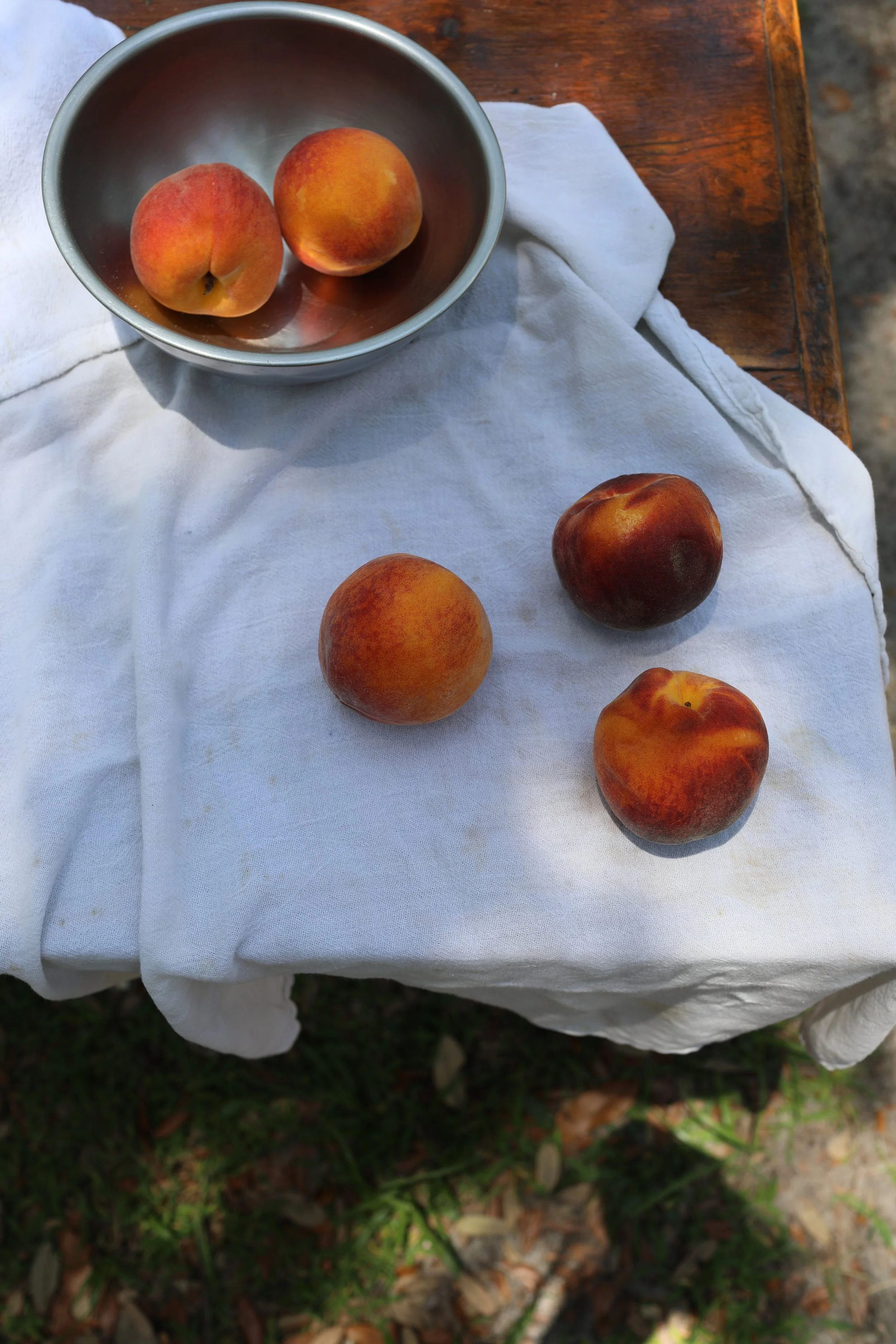 Four peaches placed on a white cloth and a metal bowl containing two peaches on a wooden surface.