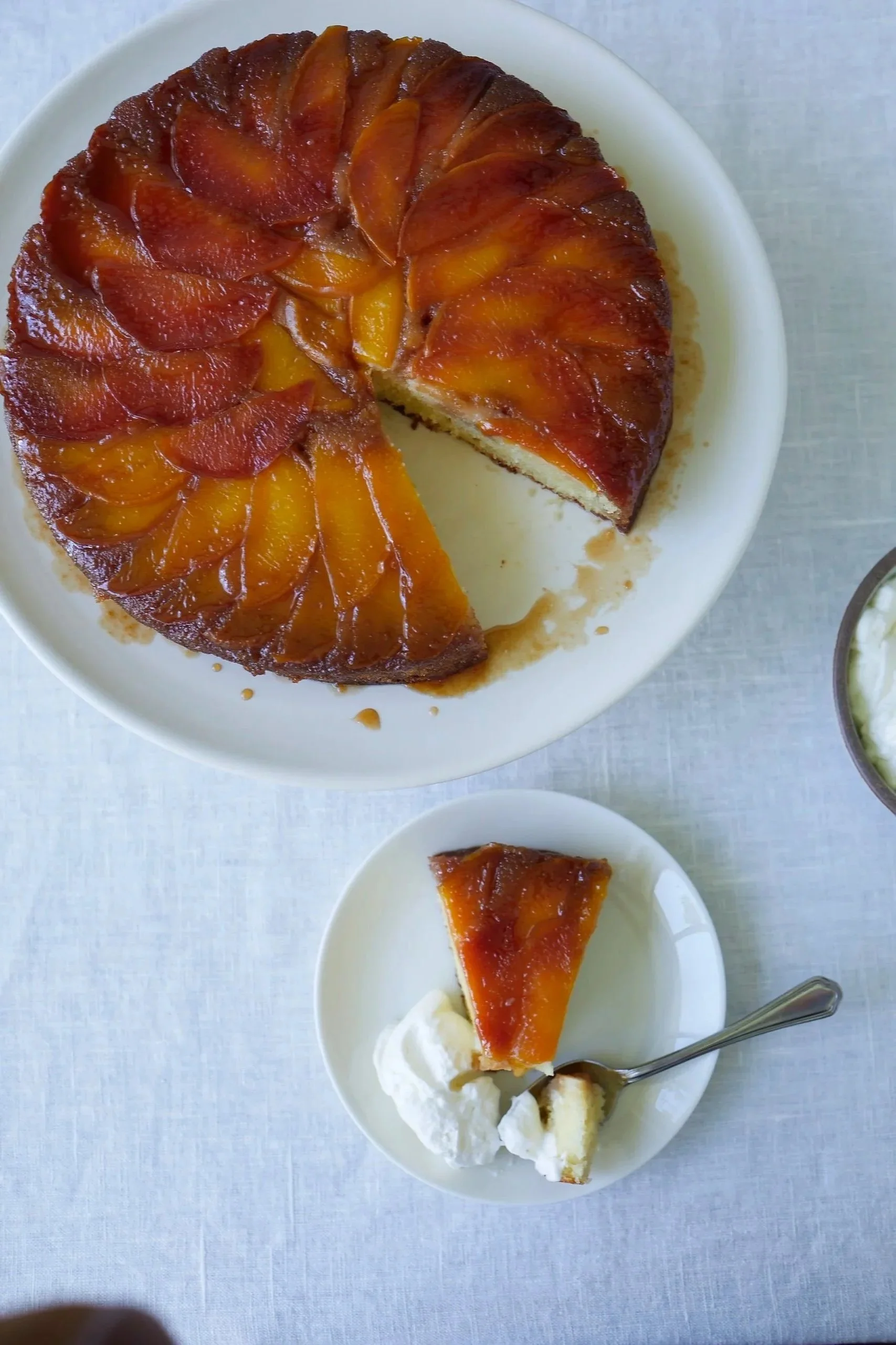 Peach upside-down cake on a white plate with a slice served on a small white dish with whipped cream, a fork, and a separate bowl of whipped cream visible.