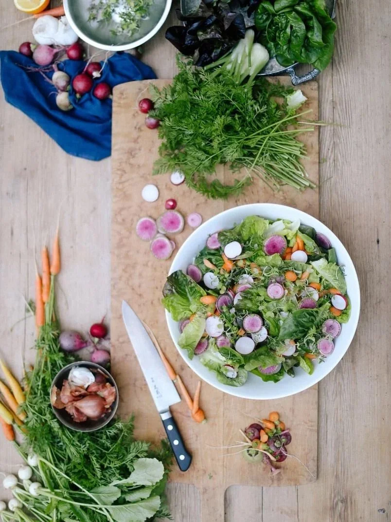 Fresh garden vegetables including radishes, carrots, green onions, and herbs on a wooden table with a bowl of mixed leafy salad and a chopping board.