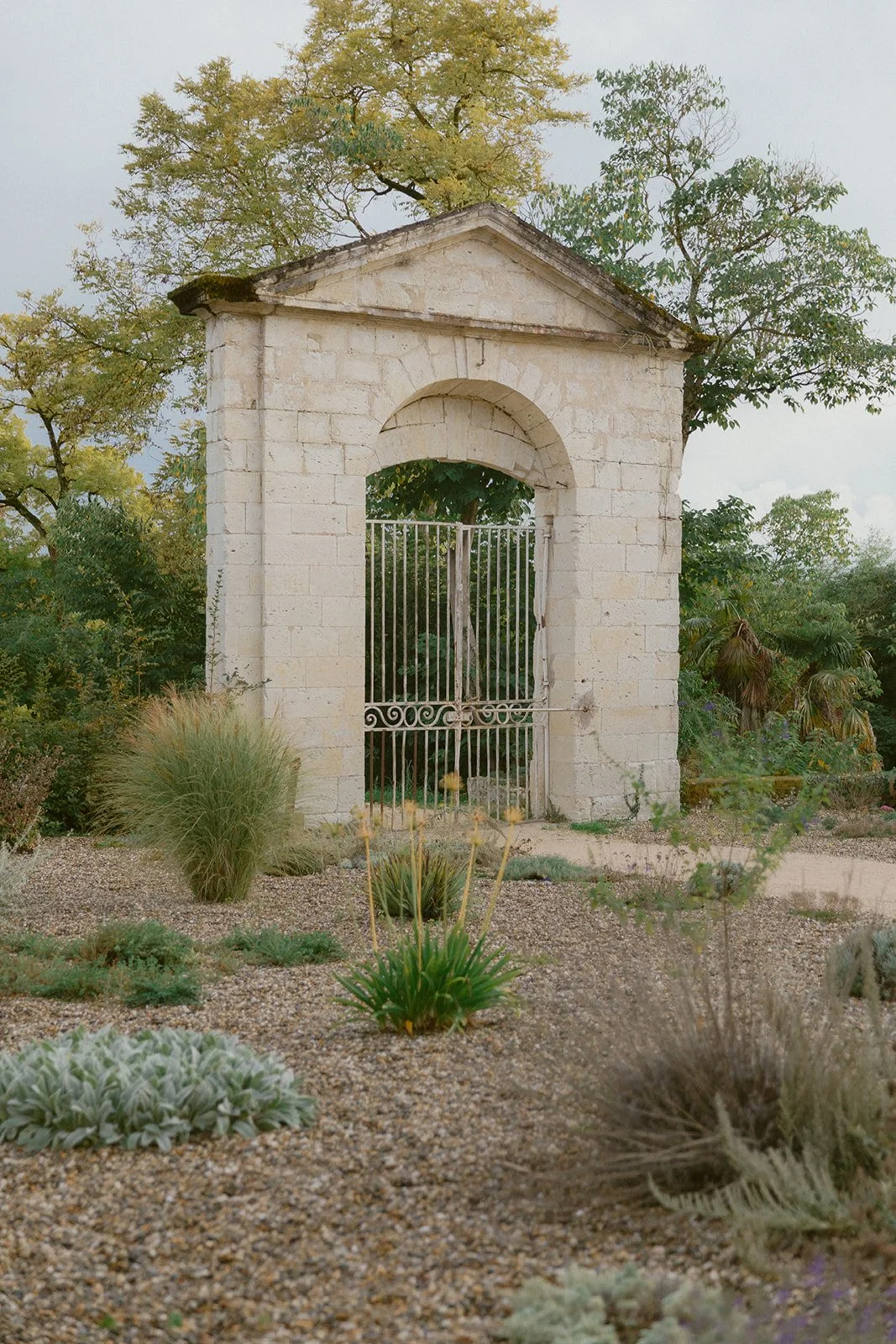 An old stone archway with a metal gate, surrounded by desert plants and trees in a garden setting.