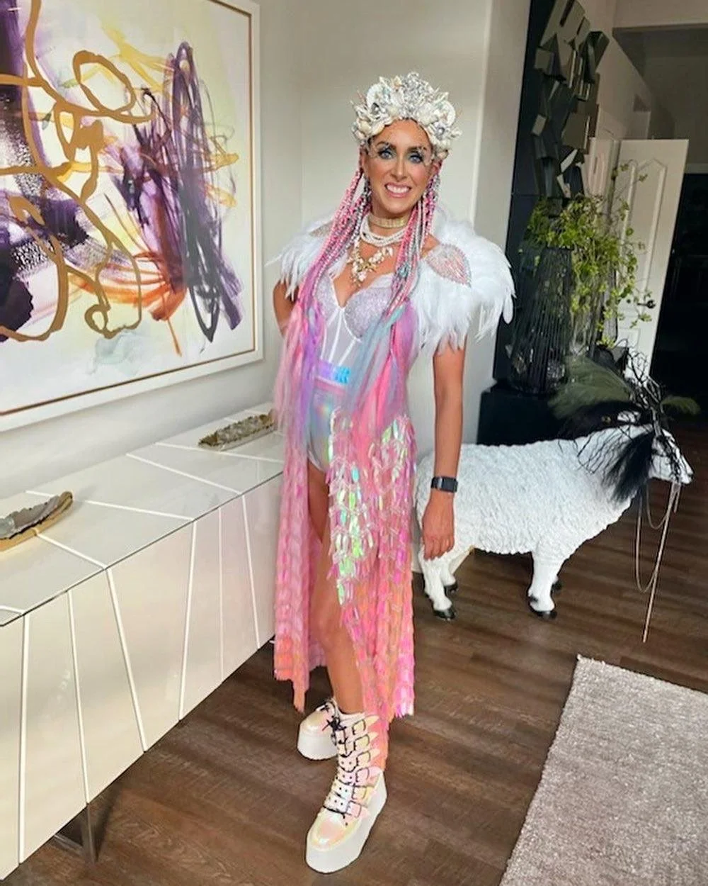 Person in colorful, iridescent outfit with feathered shoulder pieces and ornate headpiece standing indoors next to abstract painting and decorative white sheep figure.