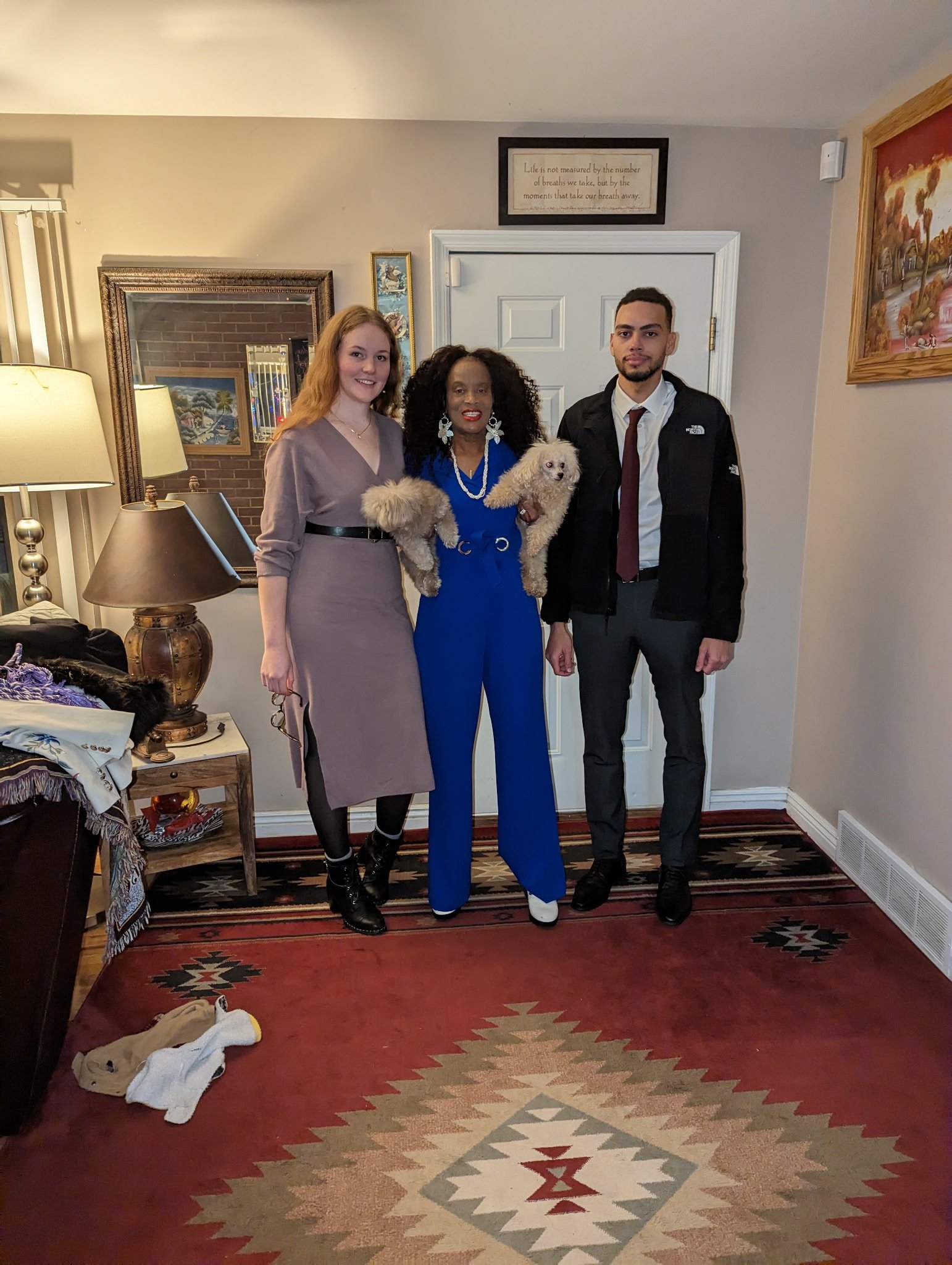 Three people standing in a living room, two are holding small fluffy dogs. A woman in a dress, a woman in a jumpsuit, and a man in a suit stand together, smiling. The room has a patterned rug, a lamp on a side table, and wall art hung around.