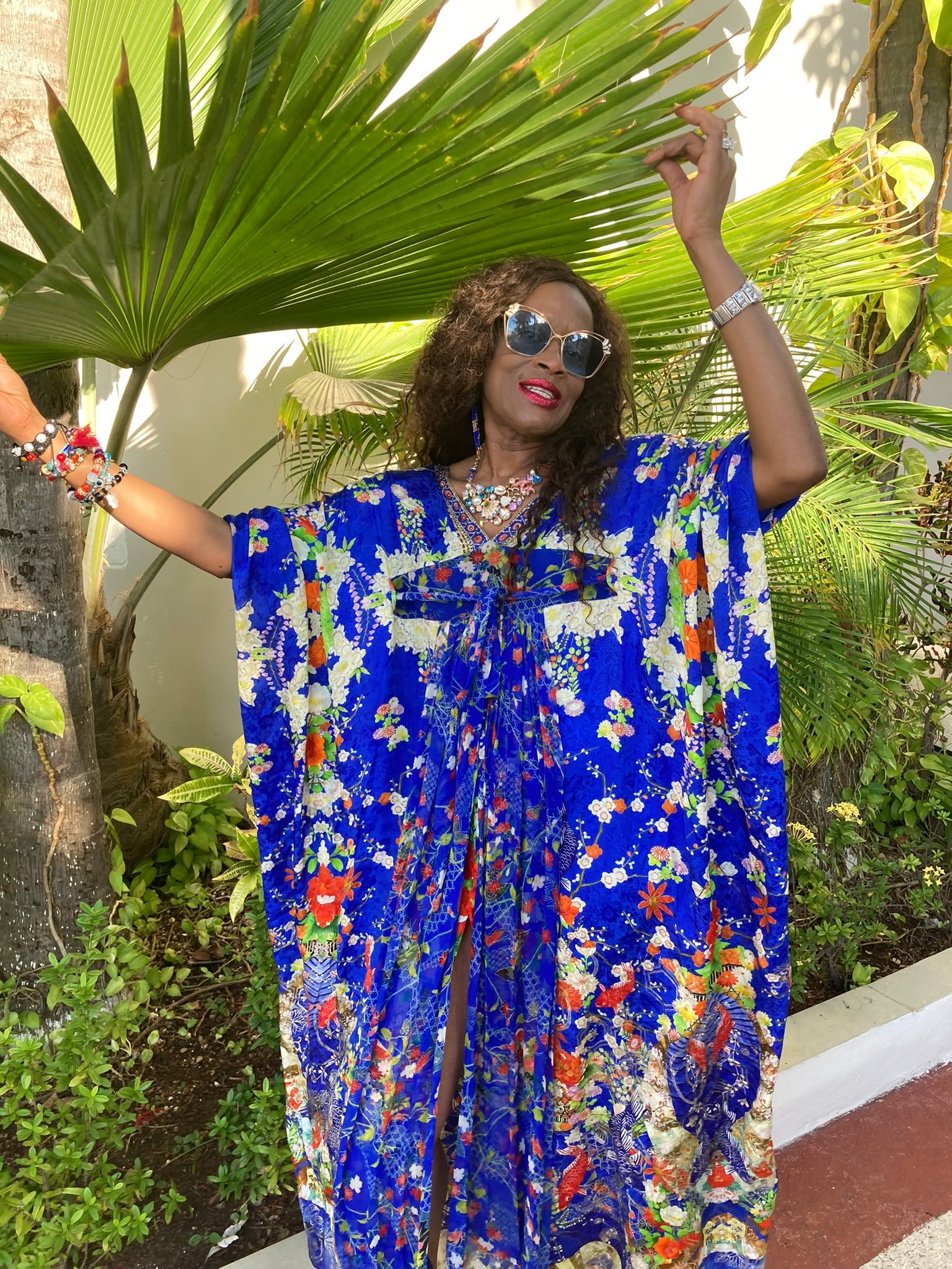 Person posing outdoors in colorful floral dress and sunglasses, surrounded by palm leaves.